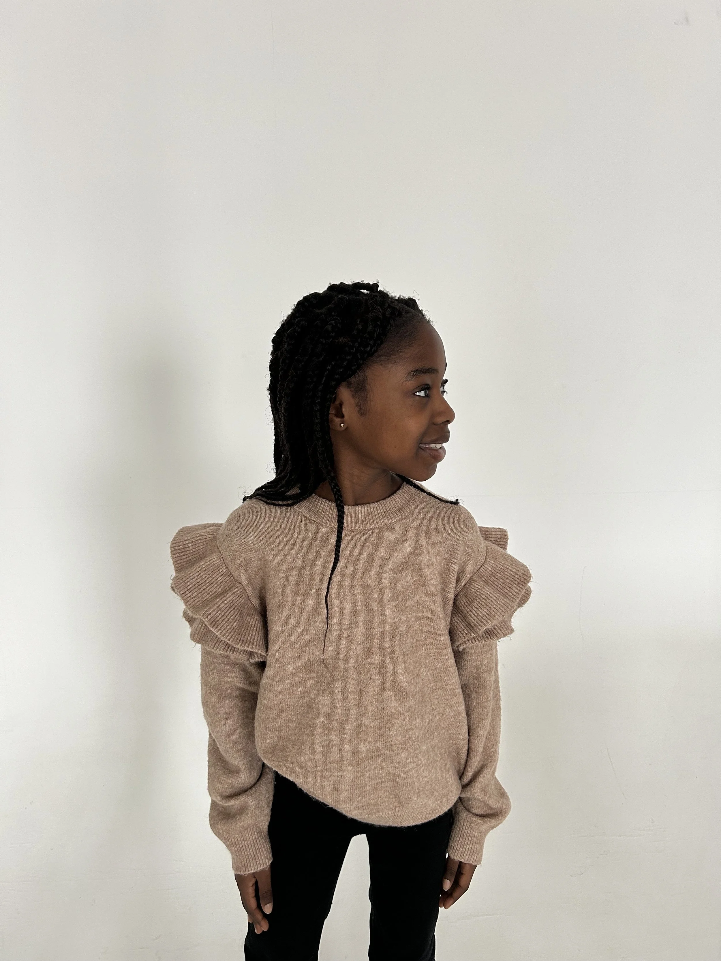 A young girl with dark braided hair wearing a tan sweater with ruffled shoulders and black pants, standing against a plain white wall, facing sideways.