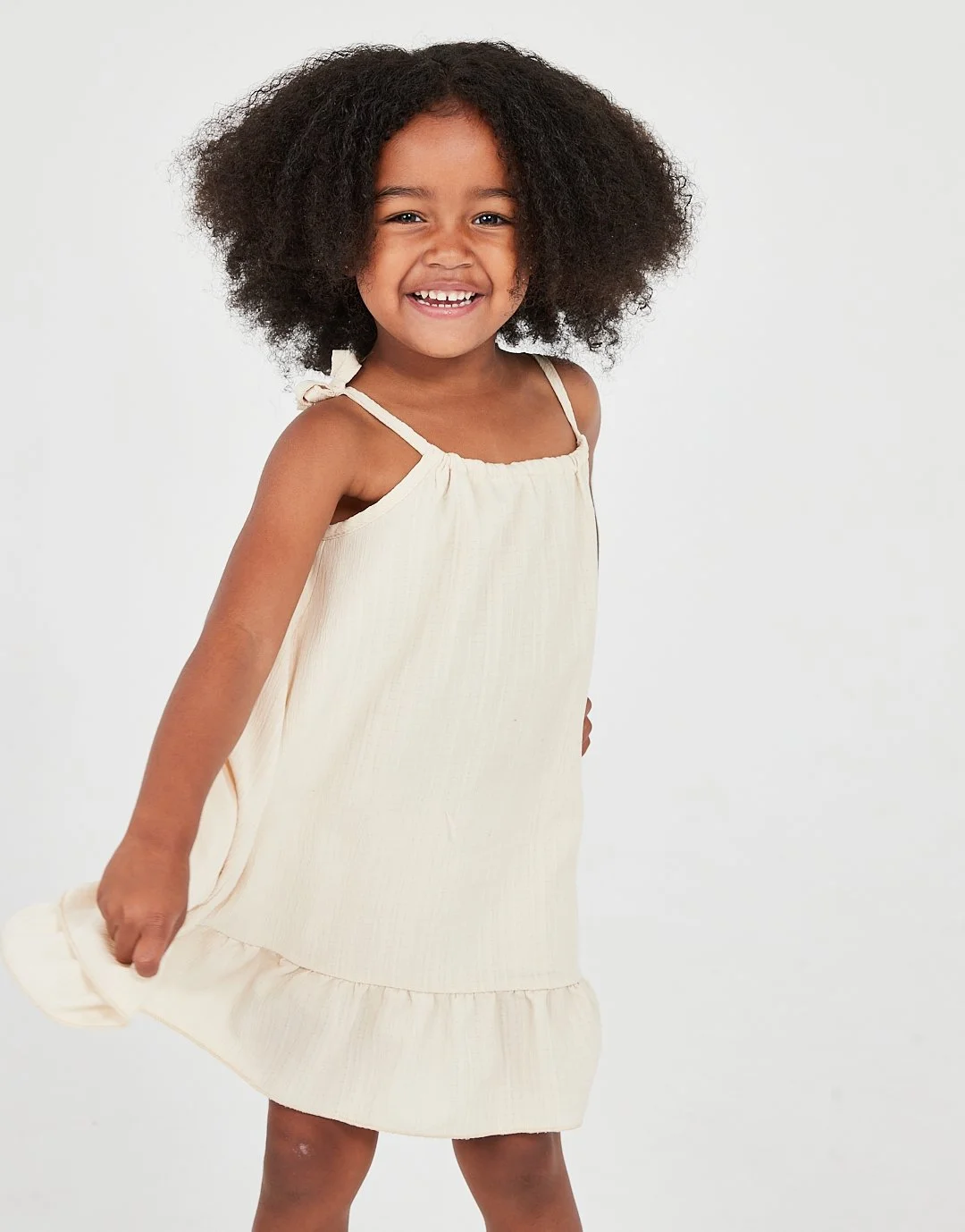 Smiling young girl with curly hair wearing a sleeveless cream-colored dress with bow straps, standing against a plain white background. Model for leading Manchester and London based child model agency Socityuk.