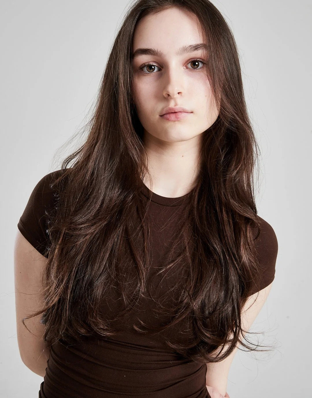 A young woman with long brown hair wearing a dark brown shirt, looking directly at the camera against a plain light grey background.