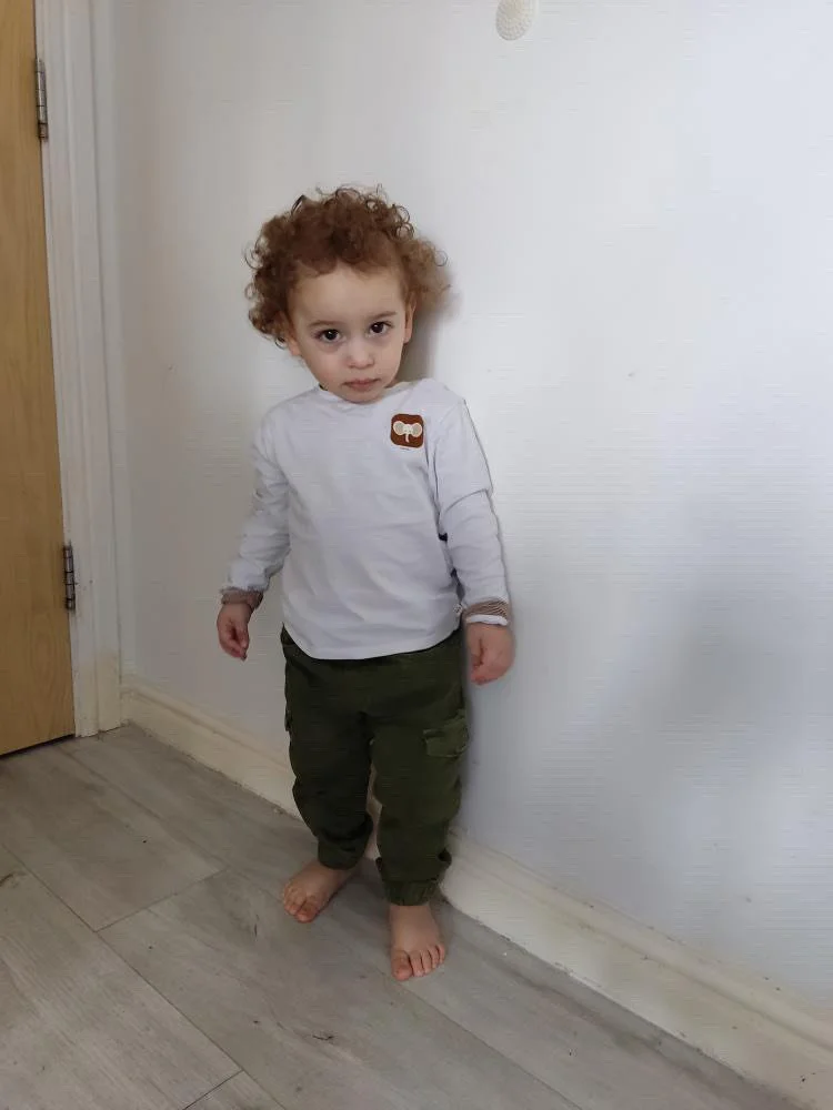A young child with curly red hair standing barefoot against a white wall, wearing a white long-sleeve shirt and green pants.