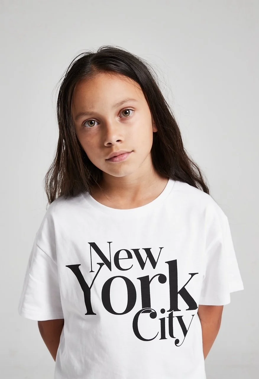 A young girl with long dark hair and light skin wearing a white T-shirt that says "New York City" in black letters, standing in front of a plain white background. Modelling for leading child model and talent agency SocityUK based in London and Manche