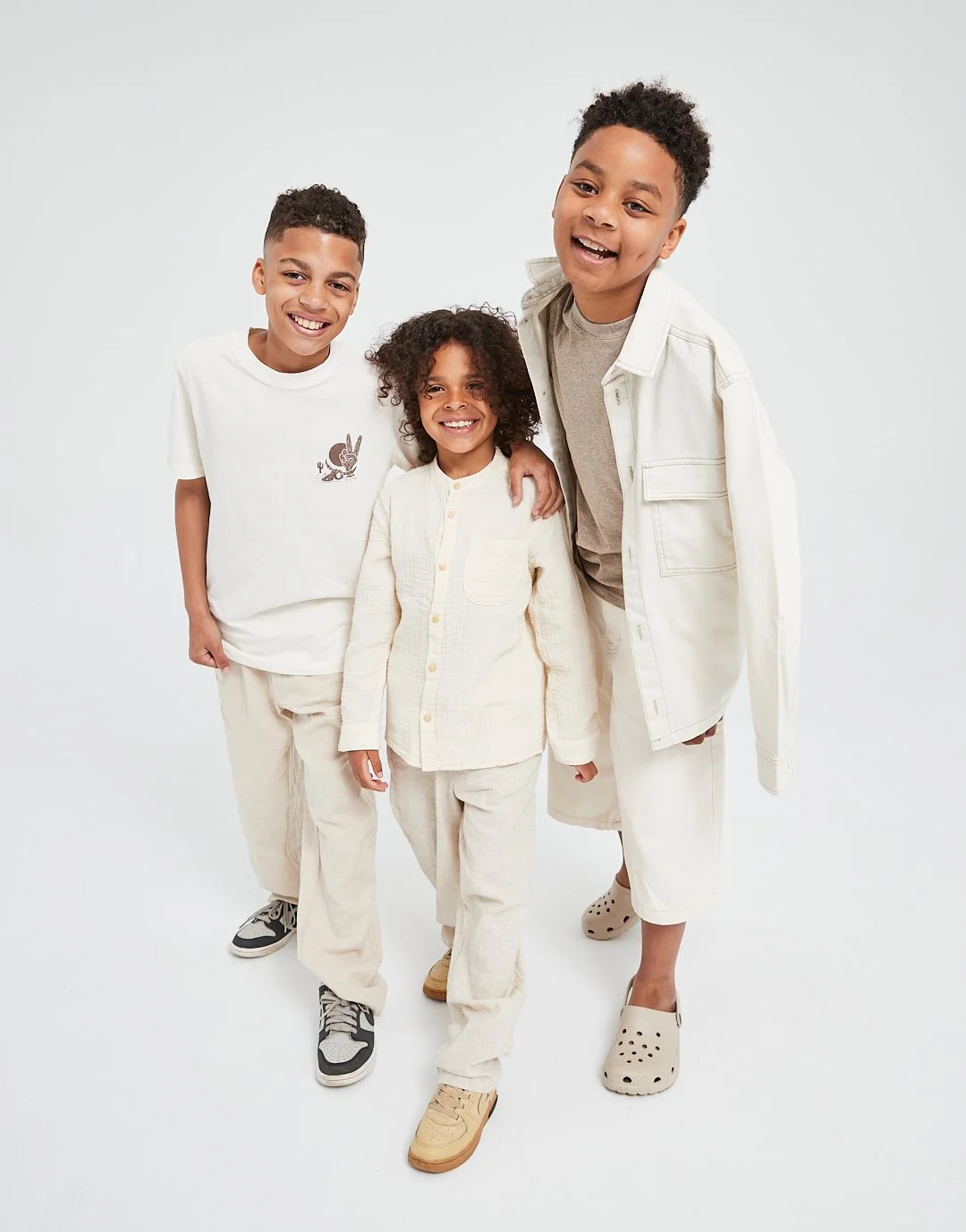 Four children smiling together in neutral-colored clothing against a plain white background.