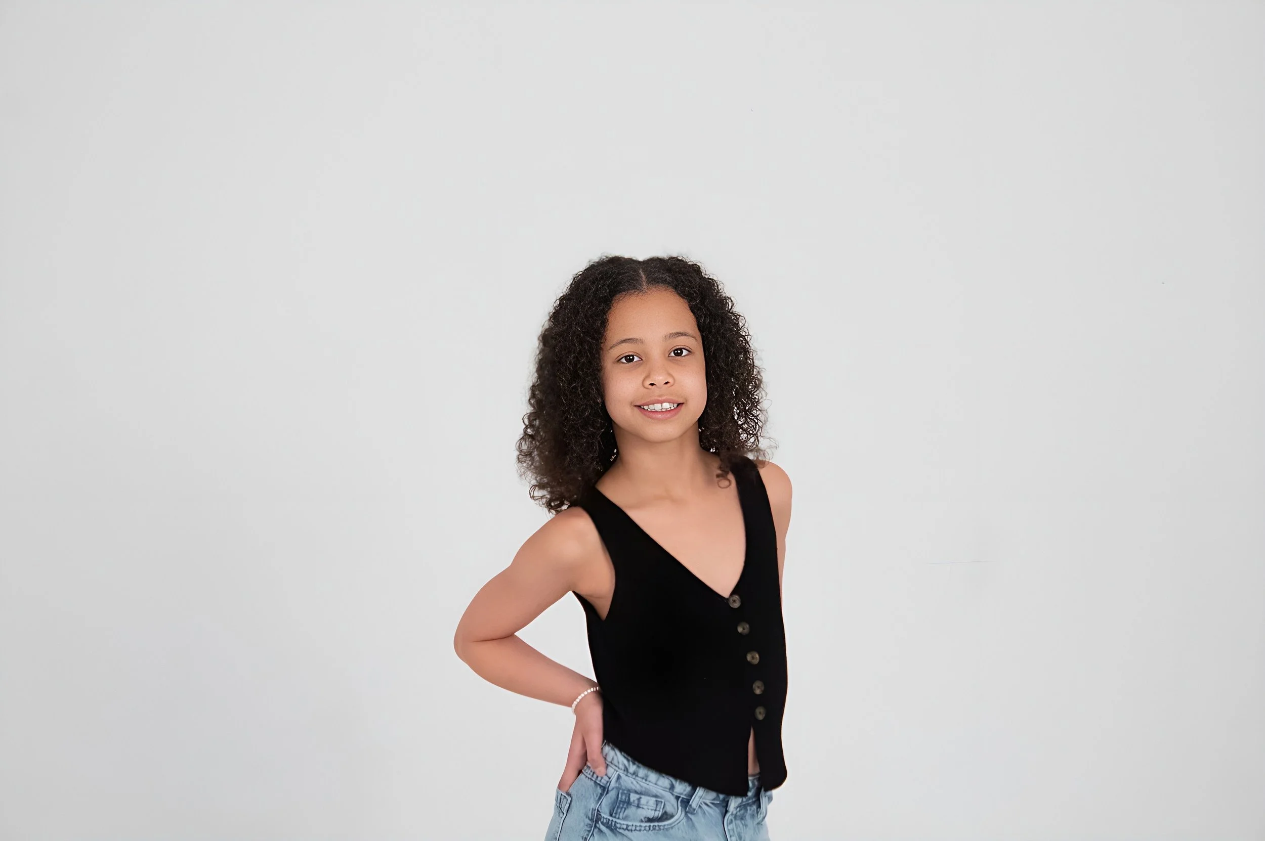 Young girl with curly hair, wearing a black sleeveless top with buttons and light blue jeans, standing against a plain white background, smiling at the camera.