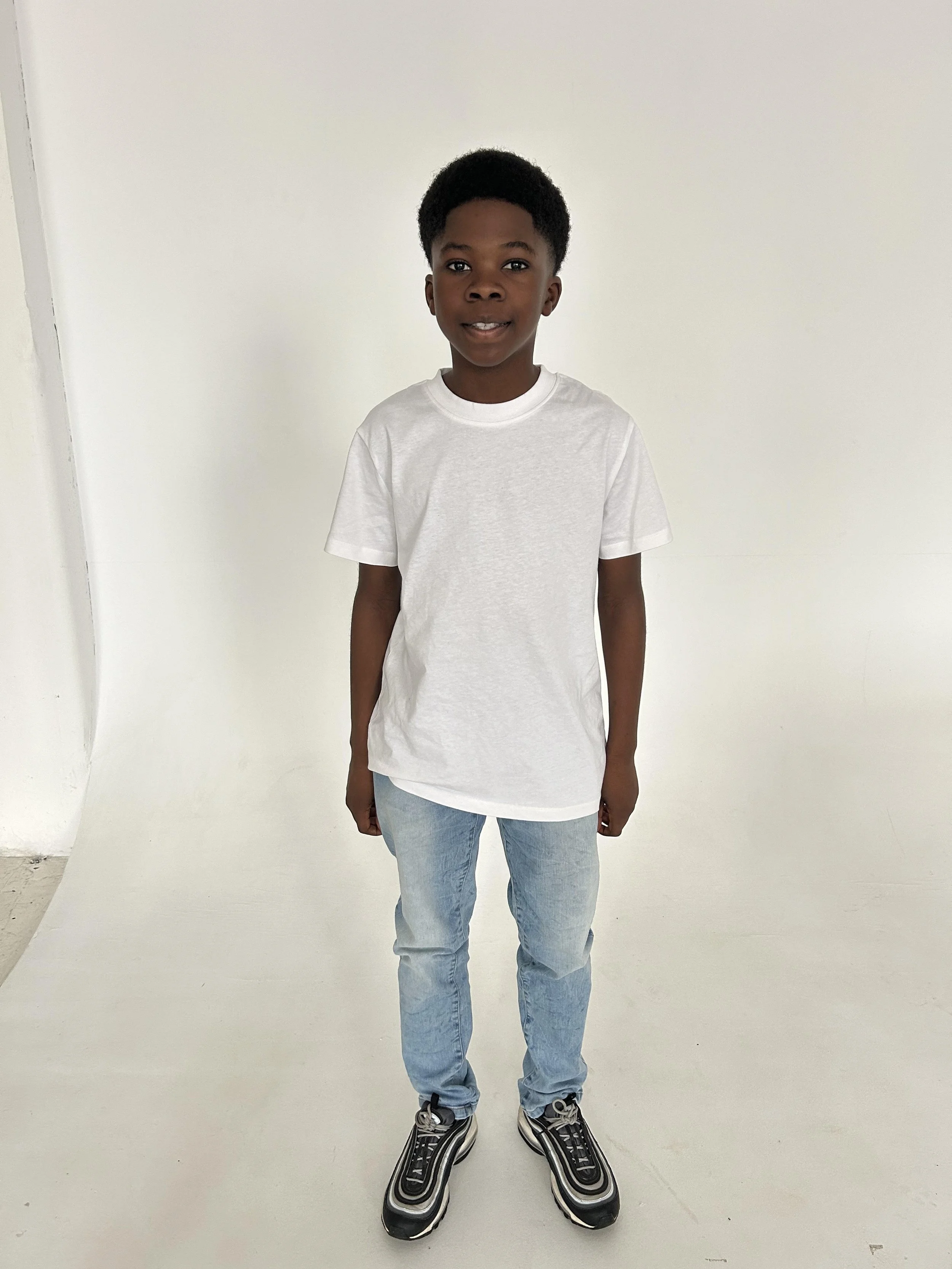 A young boy stands in front of a white background, wearing a plain white T-shirt, light blue jeans, and black sneakers.
