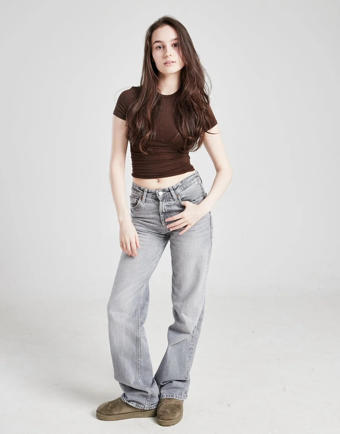 Young woman with long brown hair wearing a fitted brown crop top, loose gray jeans, and brown shoes standing against a plain light gray background.