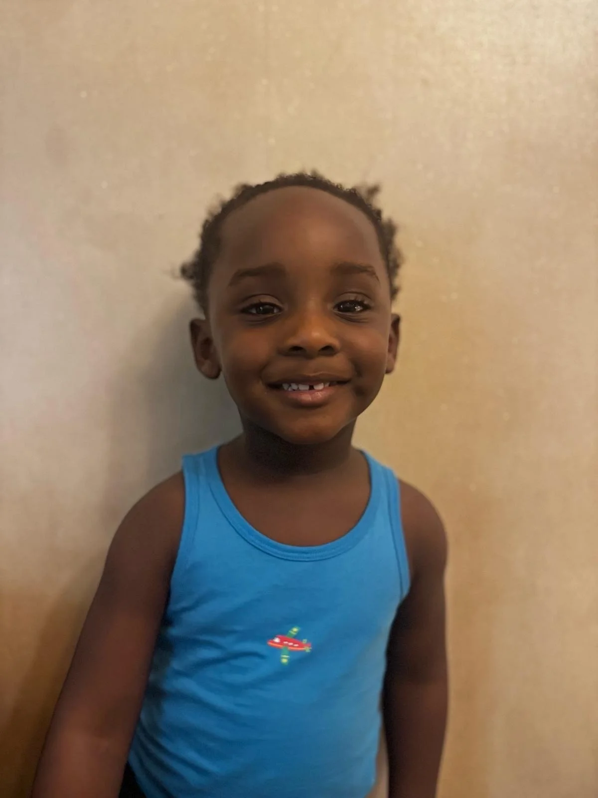Smiling young boy wearing a blue sleeveless shirt standing against a beige wall. Modelling for leading child model and talent agency SocityUK based in London and Manchester.