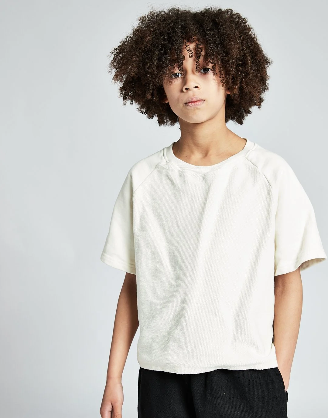 Portrait of a young person with curly hair, wearing a white t-shirt and black pants, standing against a plain, light background.