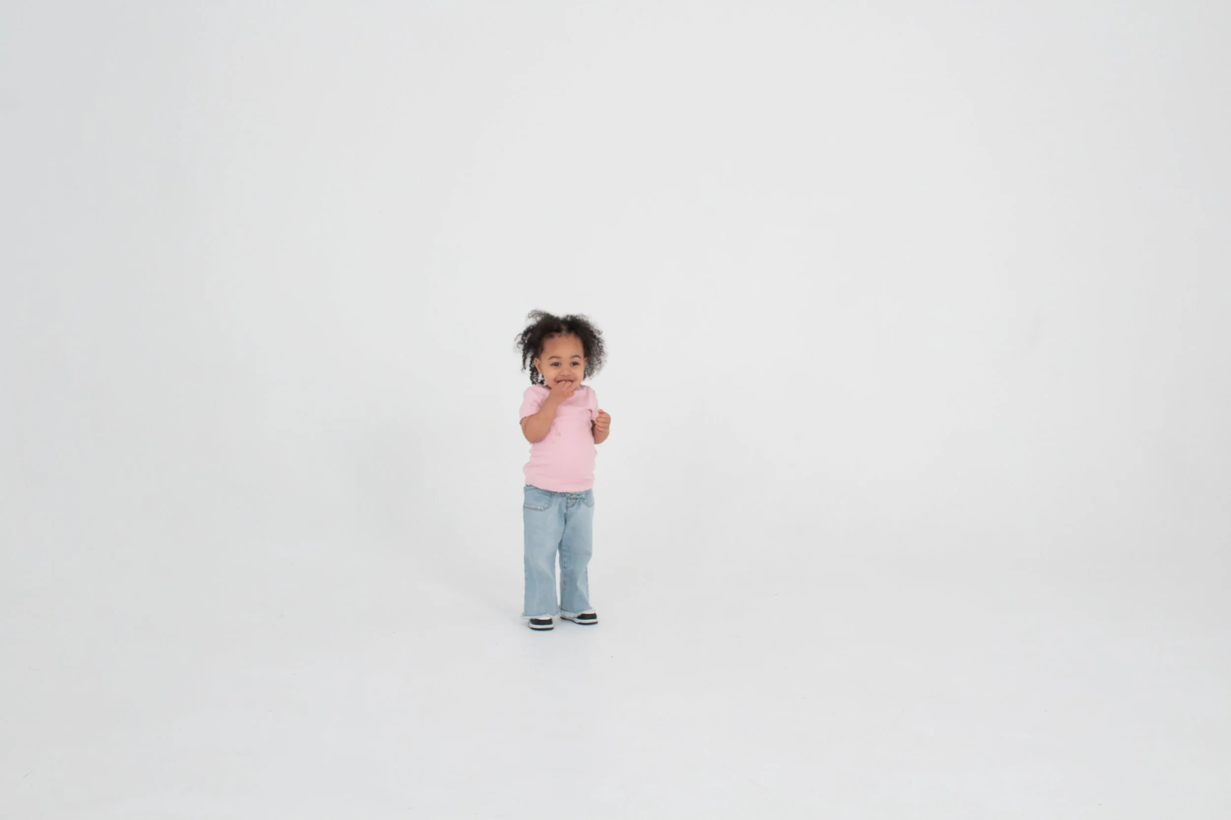 A young girl with curly hair, wearing a pink shirt, jeans, and sneakers, standing in a white space, smiling and covering her mouth with her hand.