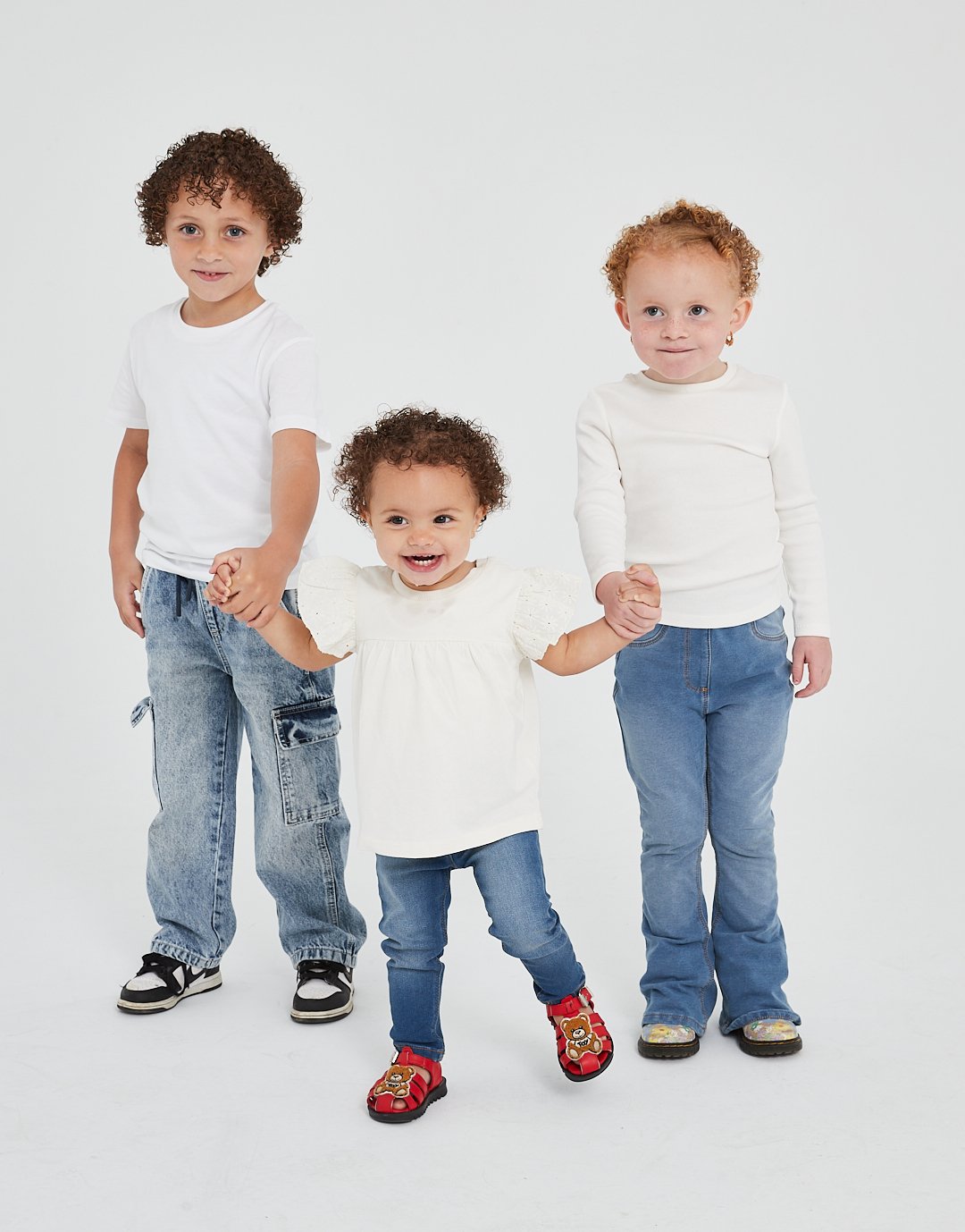 Three young children, two boys and one girl, holding hands and smiling, standing against a plain white background. The youngest girl is in the center, wearing a white top and red sandals, while the older children are wearing jeans and white tops.Mode