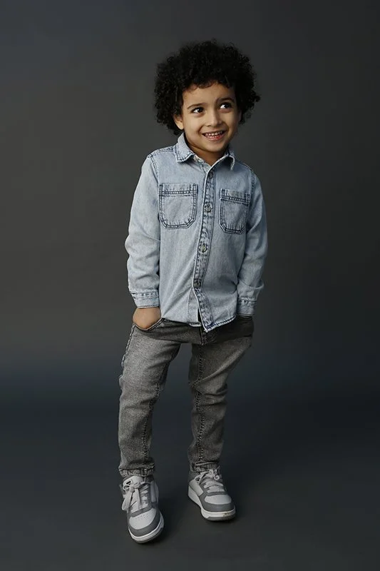 A young boy with curly hair wearing a denim shirt, gray jeans, and sneakers, smiling and standing against a dark gray background.