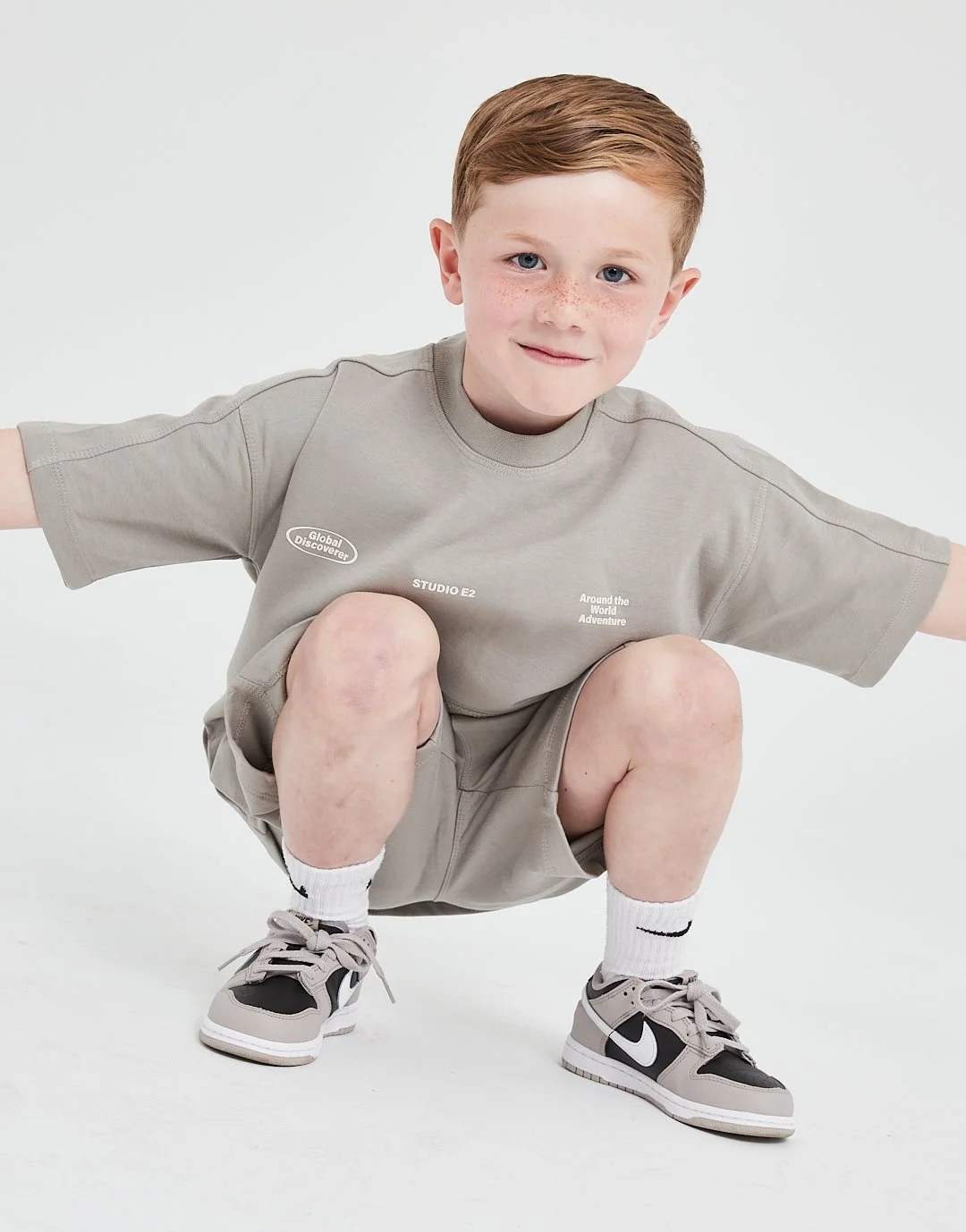 A young boy with red hair and freckles squatting with arms spread, wearing a light gray t-shirt, beige shorts, white Nike socks, and gray and black Nike sneakers, against a plain white background.