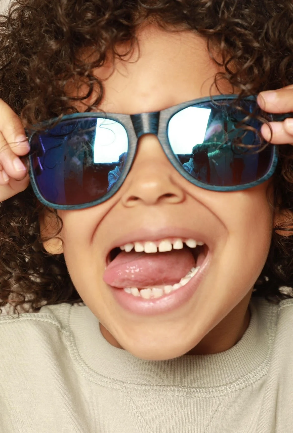 A young child with curly hair wearing reflective sunglasses, smiling and sticking out tongue, reflections of a window and person are visible in the sunglasses.