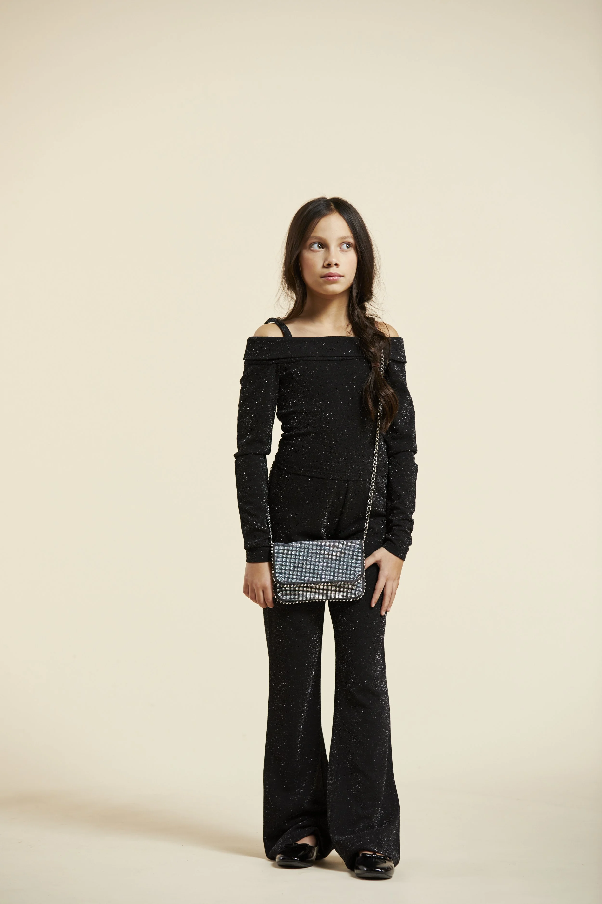 Young woman with long dark hair wearing a black off-the-shoulder top and matching flared pants, holding a sparkly silver clutch purse, standing against a plain beige background. Modelling for leading child model and talent agency SocityUK based in Lo