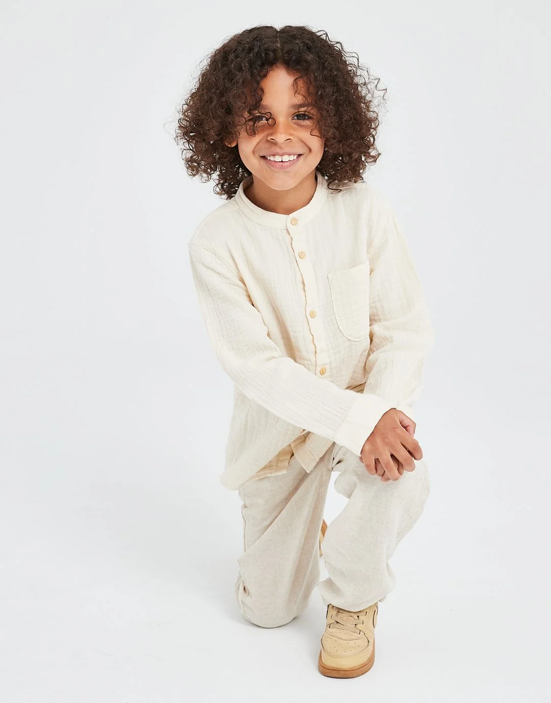 A smiling young child with curly hair, wearing a cream-colored button-up shirt and matching pants, kneeling on one knee against a plain white background.