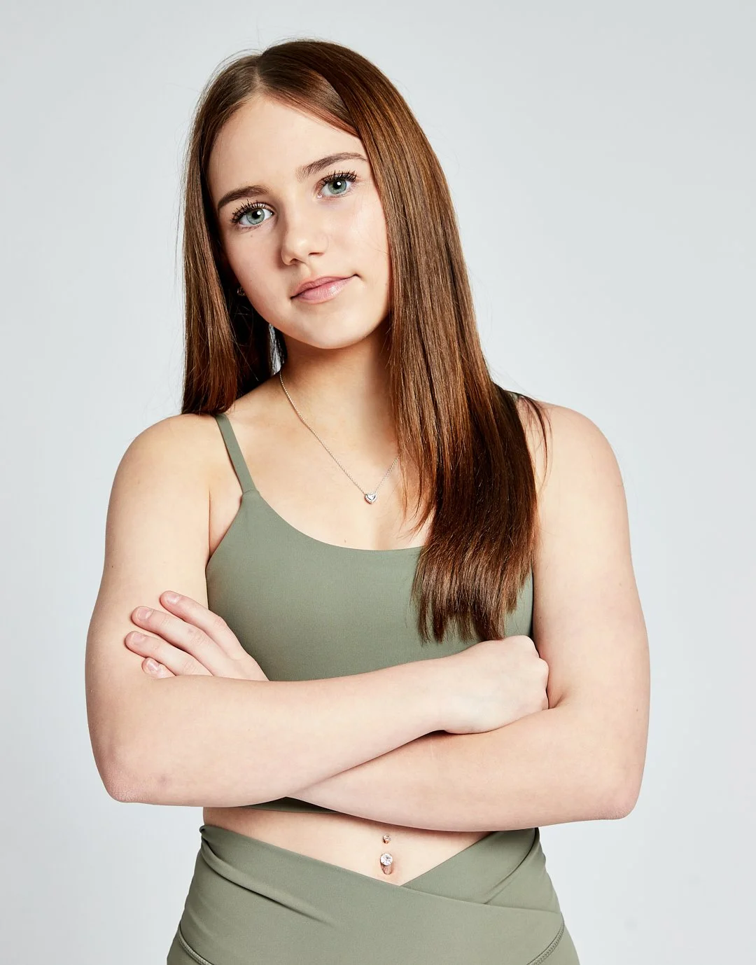 Young model with long brown hair, green eyes, wearing a green tank top, with arms crossed, standing against a plain light gray background. Modelling for leading child model and talent agency SocityUK.
