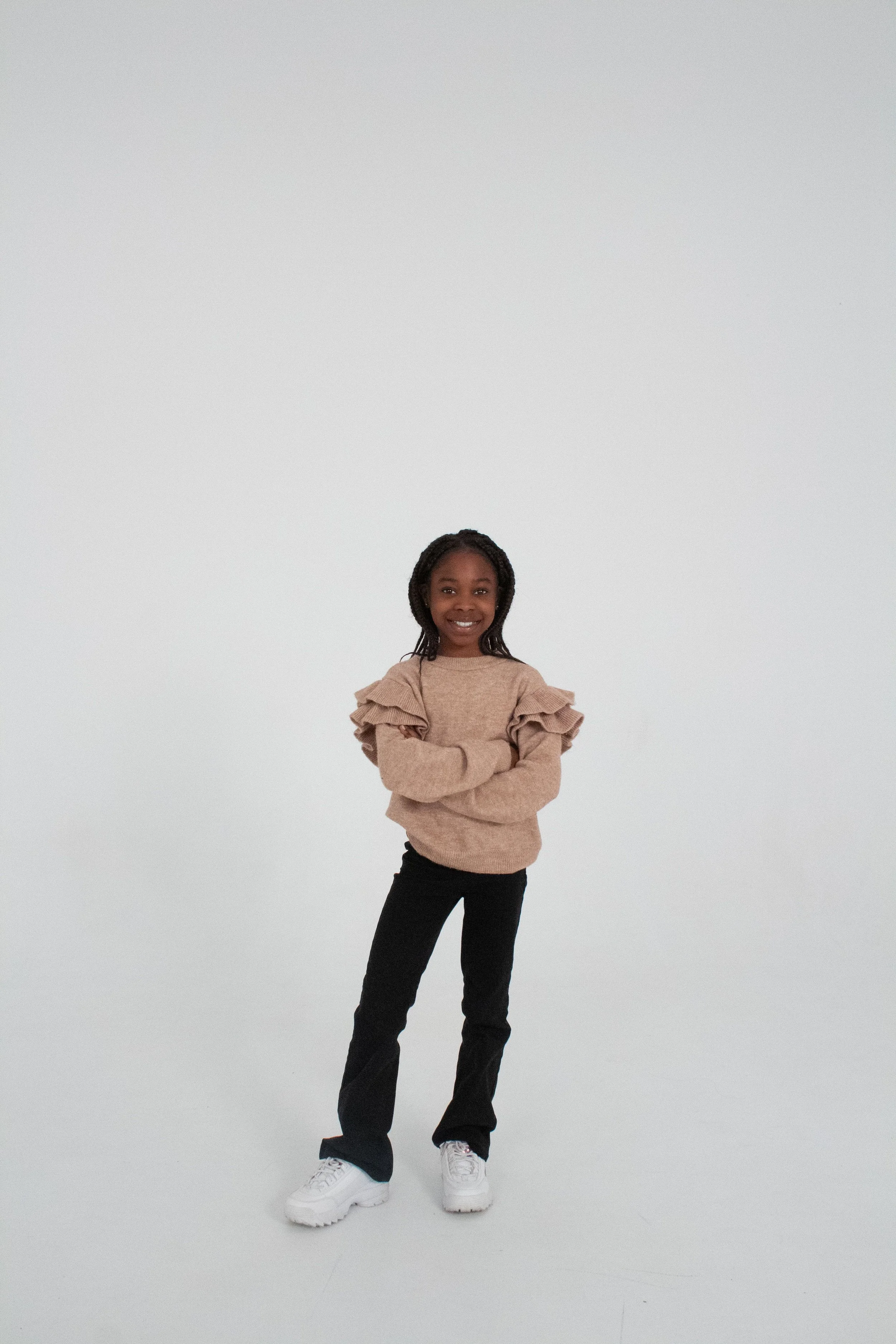 A young girl with braided hair smiling and crossing her arms, wearing a ruffled beige sweater, black pants, and white sneakers, standing against a plain white background.