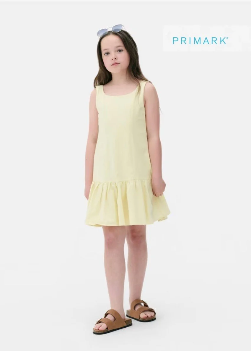 A young girl wearing a light yellow sleeveless dress with ruffle at the hem, brown sandals, and sunglasses on her head, standing against a plain white background with the Primark logo in the top right corner.