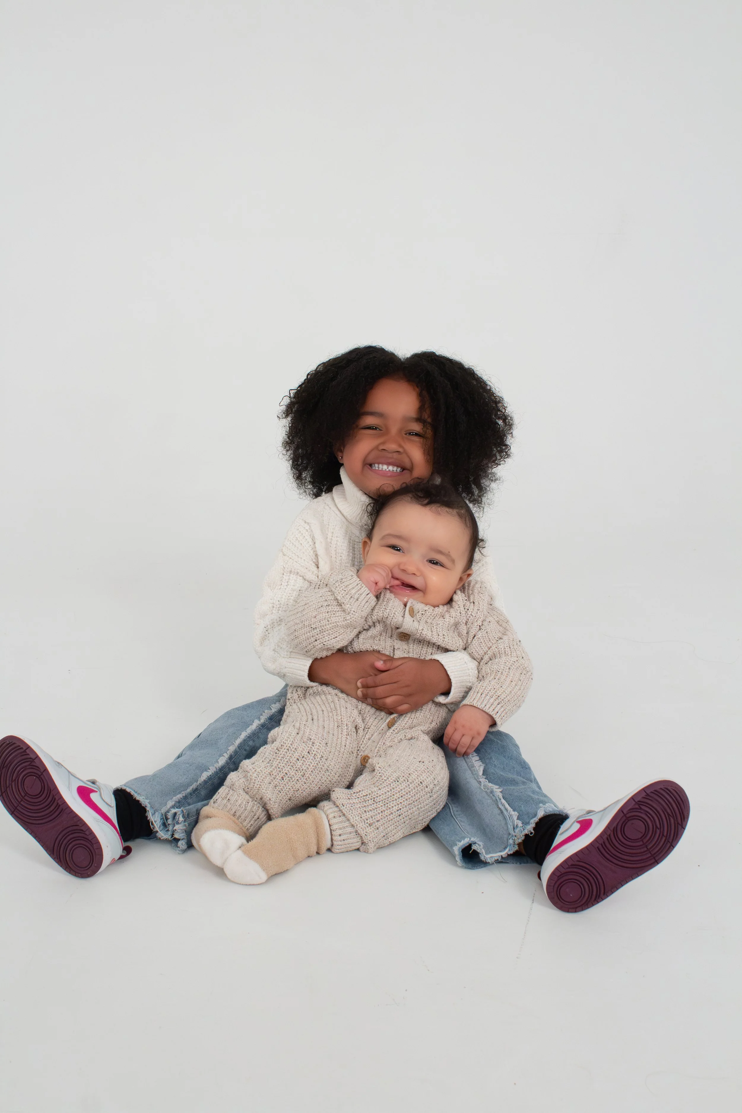 Two children, a girl and a baby boy, sitting together on the floor against a plain white background, smiling and hugging each other. Model for leading Manchester and London based child model agency Socityuk.