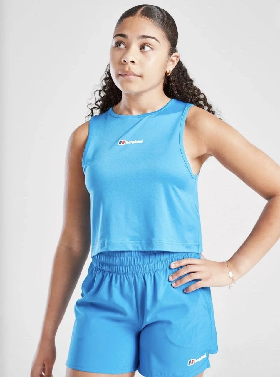 A young woman with curly hair wearing a blue sleeveless athletic top and matching shorts, standing with one hand on her hip against a plain light background.