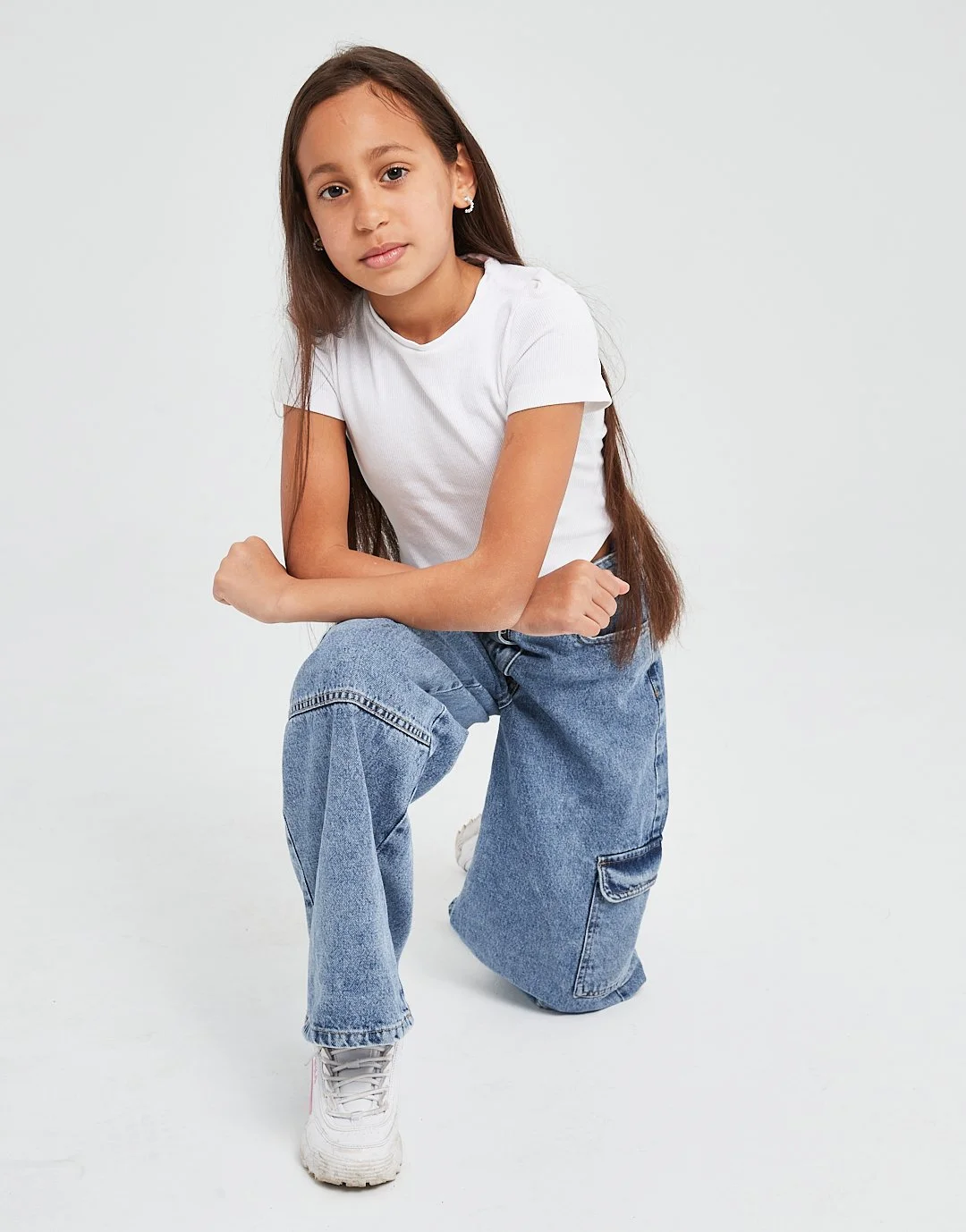 A young girl with long brown hair, wearing a white t-shirt, loose blue jeans, and white sneakers, is kneeling on one knee, looking at the camera with a neutral expression. Modelling for leading child model and talent agency SocityUK based in London a