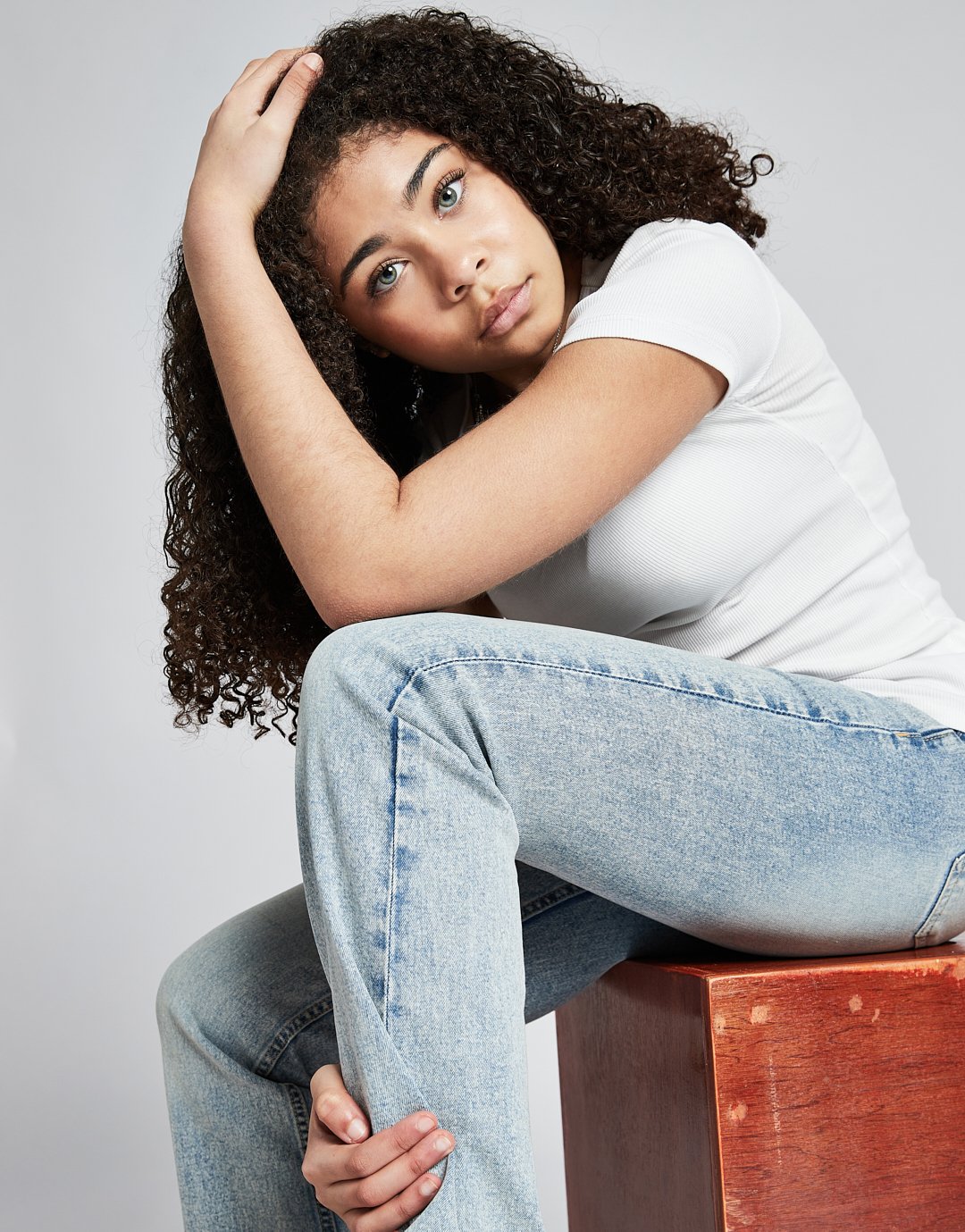 Young woman with curly hair sitting on a wooden block, wearing a white t-shirt and jeans, resting her head on her arm with a neutral expression.