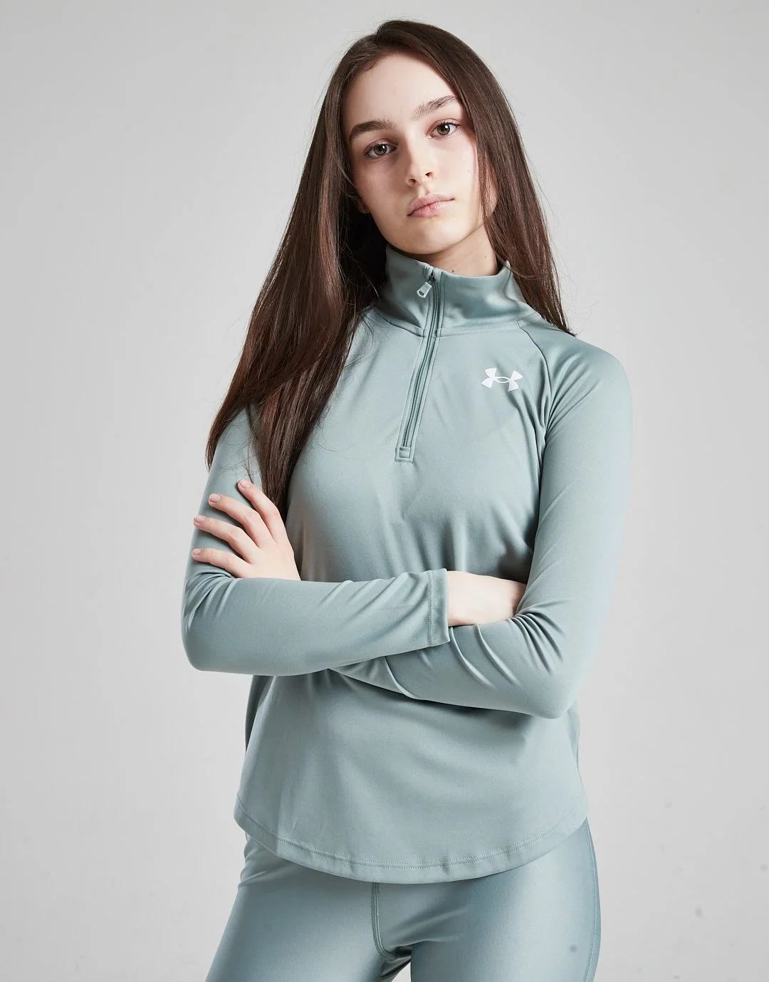 Young woman with long brown hair wearing a light gray Under Armour athletic top with a quarter-zip and matching leggings, standing with arms crossed against a plain gray background.