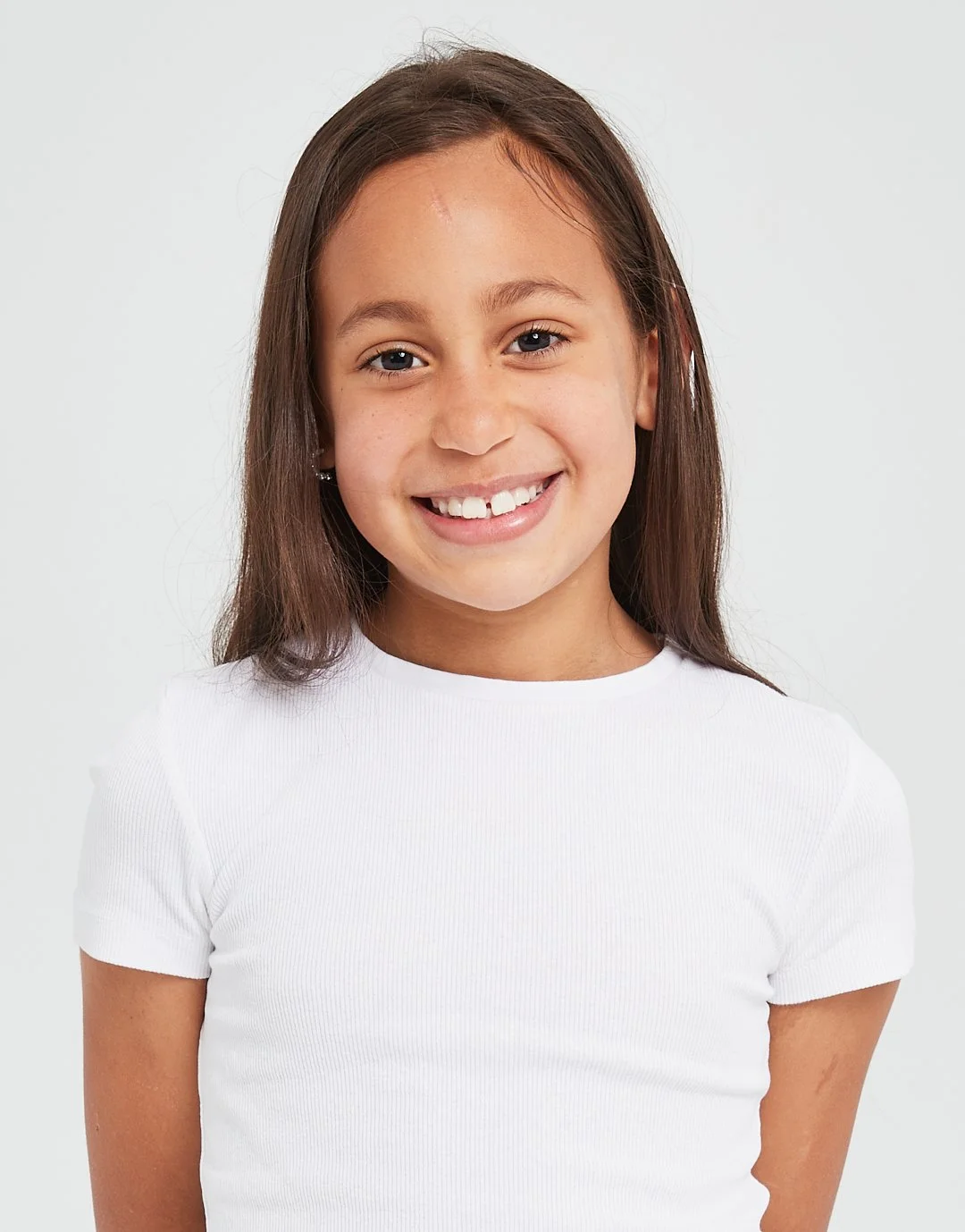 A young girl with long brown hair smiling and showing missing front teeth, wearing a white T-shirt, against a plain white background. Modelling for leading child model and talent agency SocityUK based in London and Manchester.