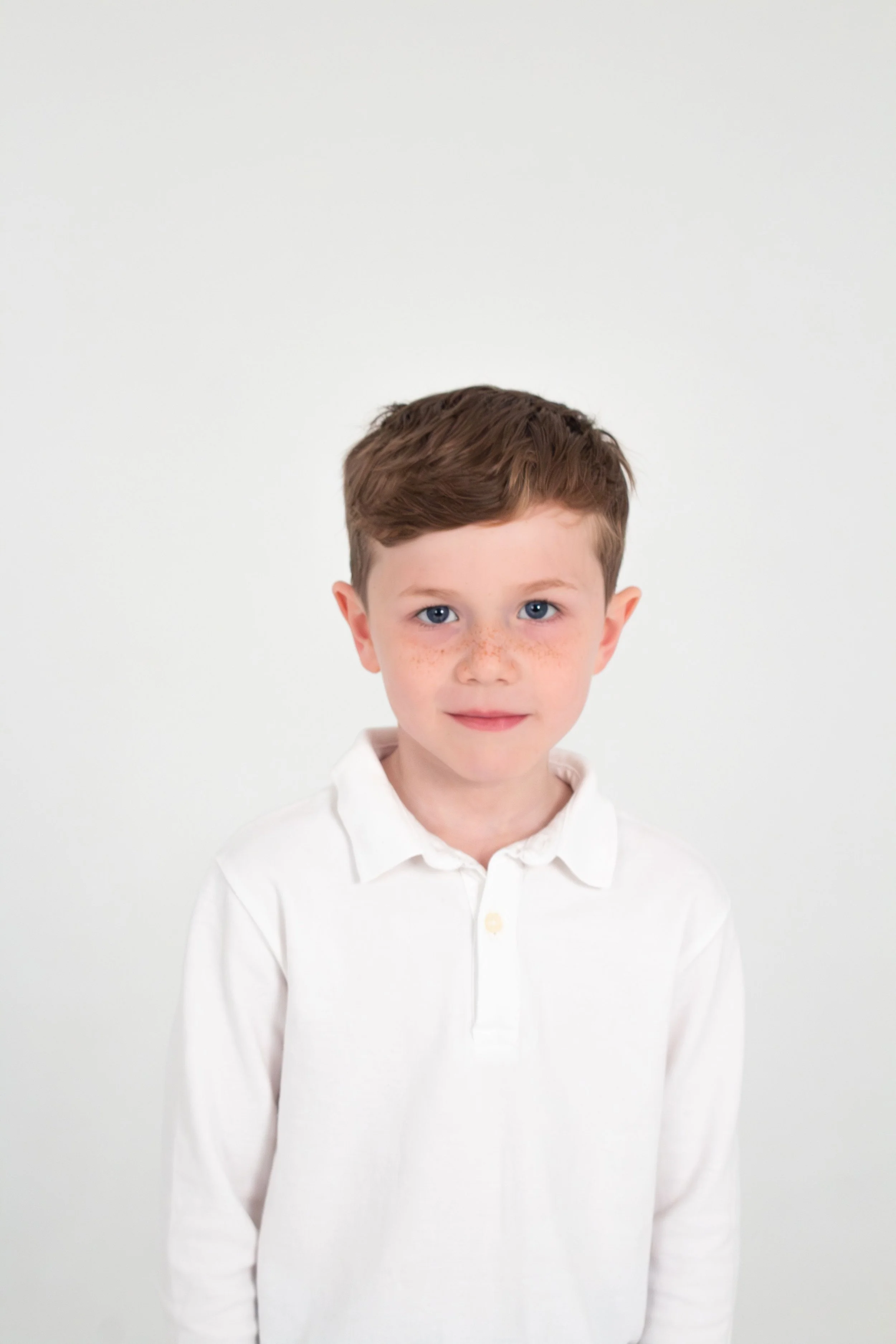 A young boy with light skin, brown hair, and blue eyes, wearing a white collared shirt, standing against a plain white background.