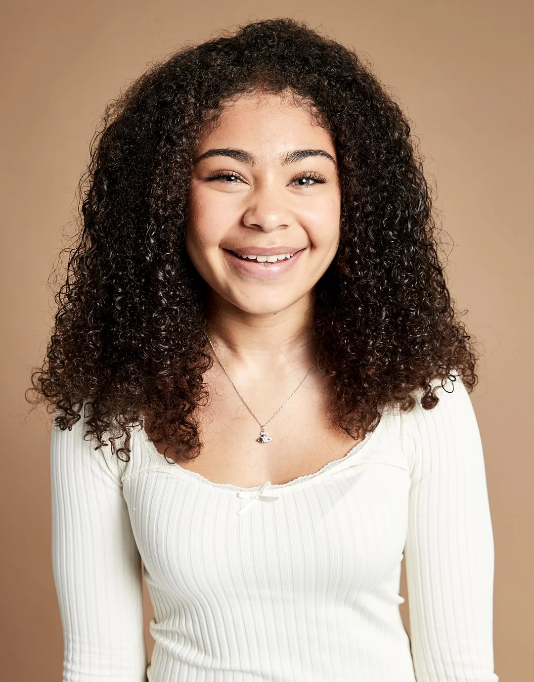 Smiling young woman with curly hair wearing a white long-sleeve top and a necklace against a neutral background.