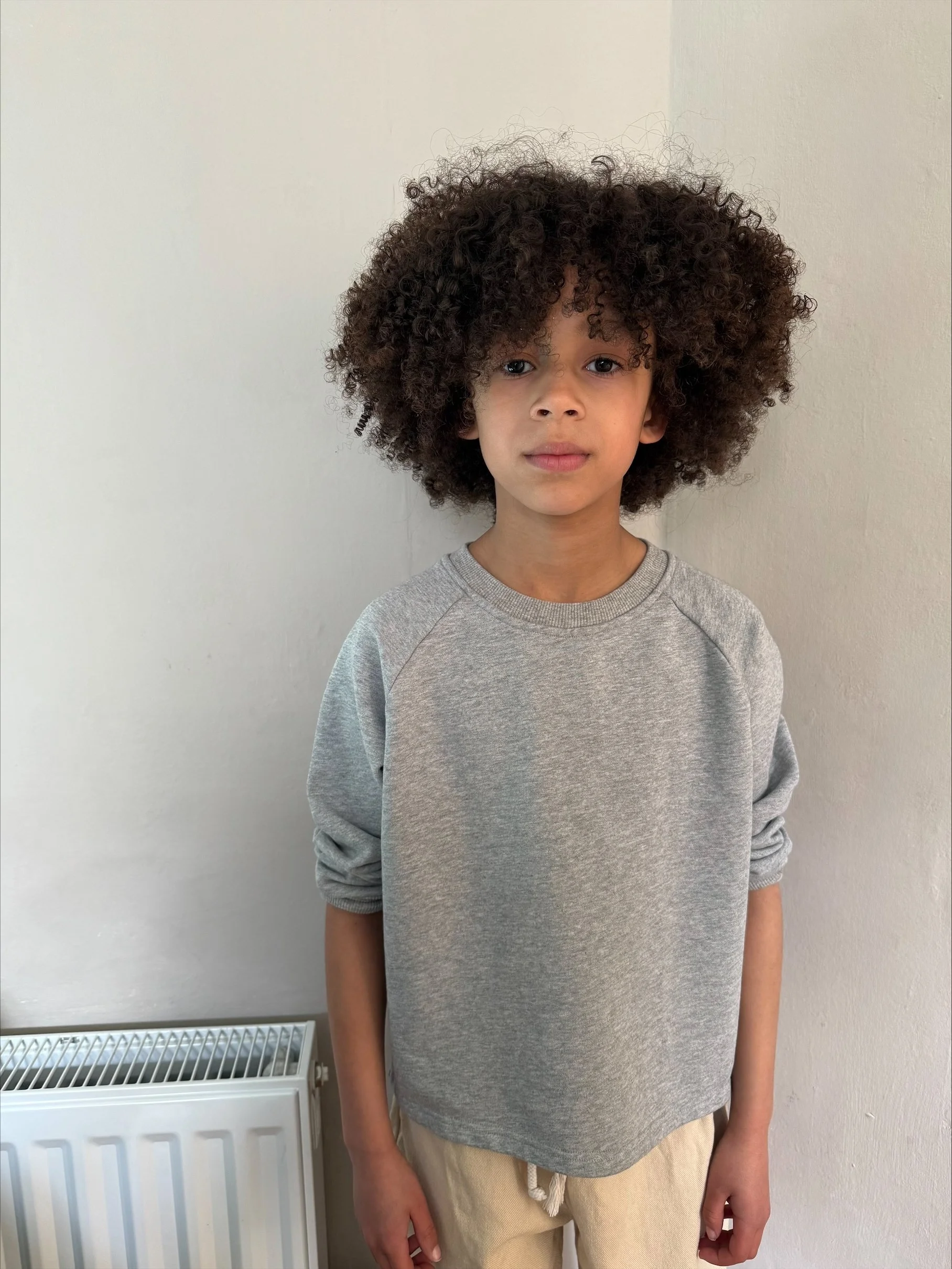 A young boy with curly dark hair and light brown skin, wearing a gray sweatshirt and beige pants, standing in front of a plain light-colored wall with a radiator at his side.