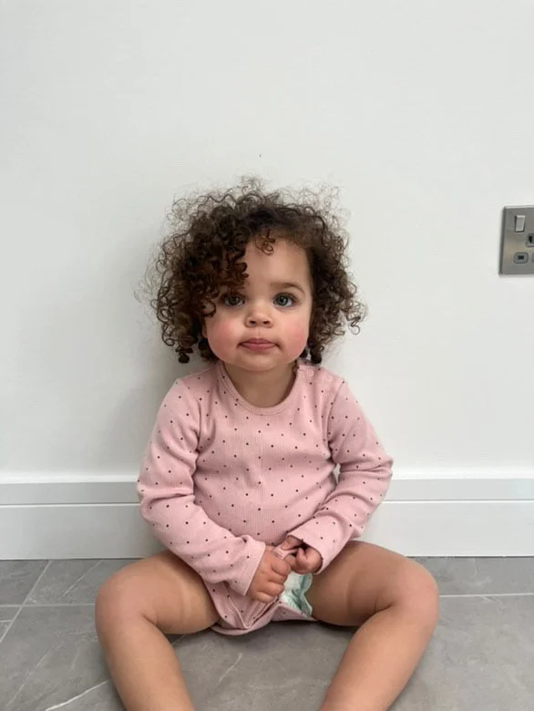 A young girl with curly hair sitting on the floor against a white wall, wearing a pink long-sleeve shirt with small dots and underwear, looking at the camera with a neutral expression. Modelling for leading child model and talent agency SocityUK base