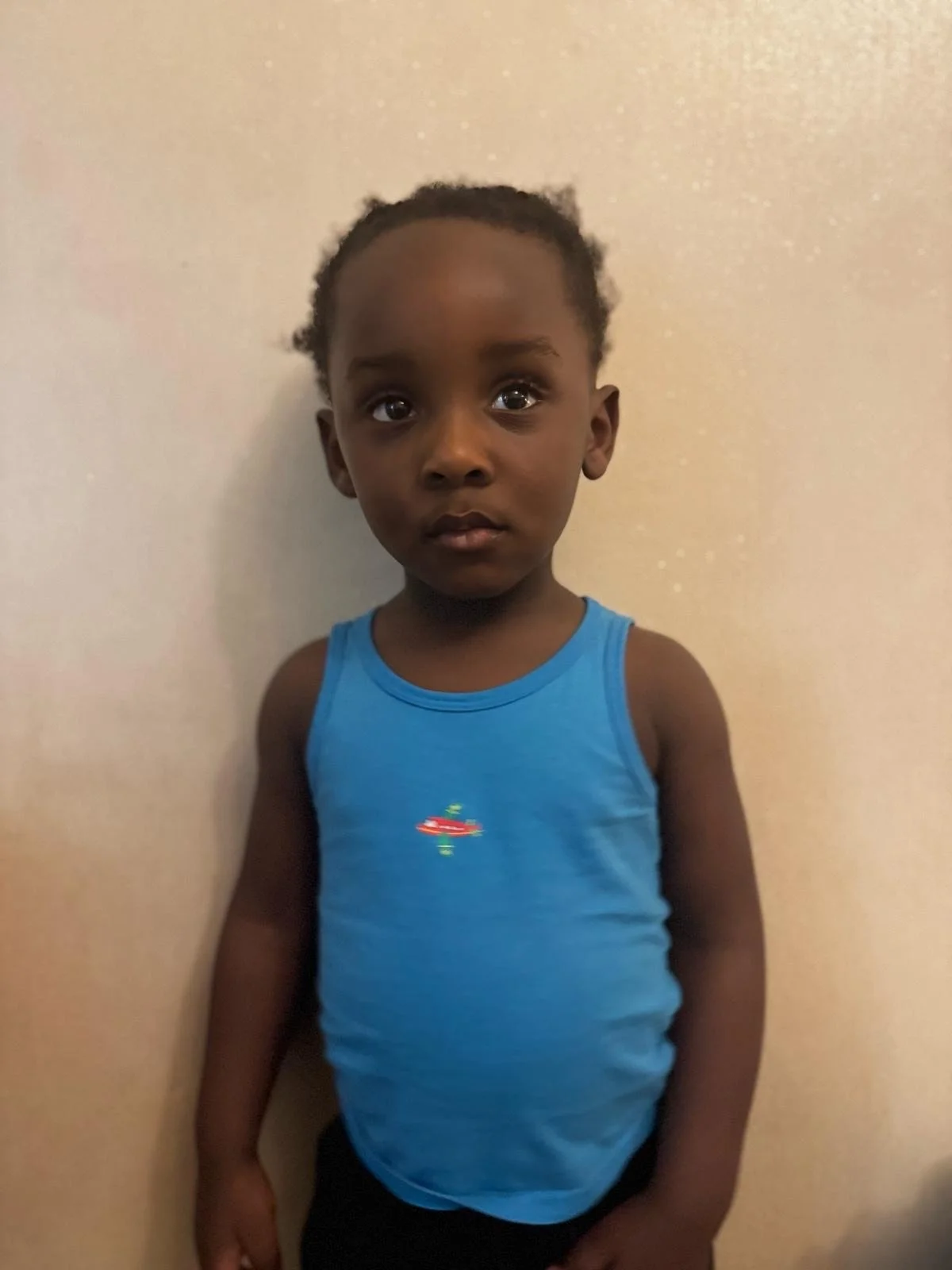 A young boy with dark hair and brown skin wearing a blue tank top stands against a plain off-white wall.Modelling for leading child model and talent agency SocityUK based in London and Manchester.