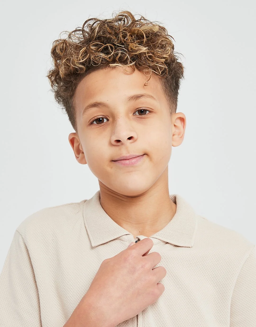 A boy with curly hair wearing a beige polo shirt, holding the zipper at his neck, looking at the camera with a slight smile.