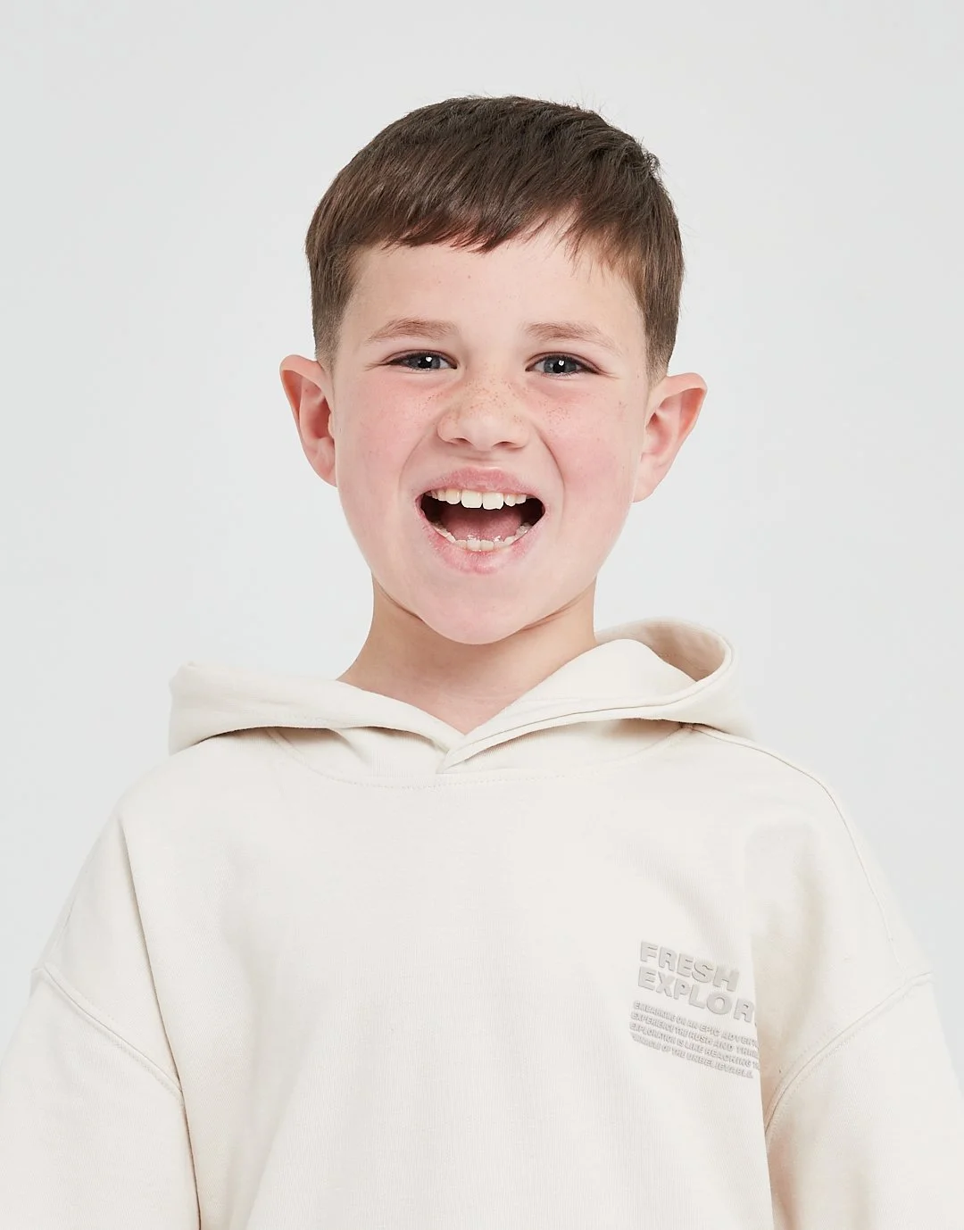 Young boy smiling with missing front teeth, wearing a white hoodie, against a plain white background.