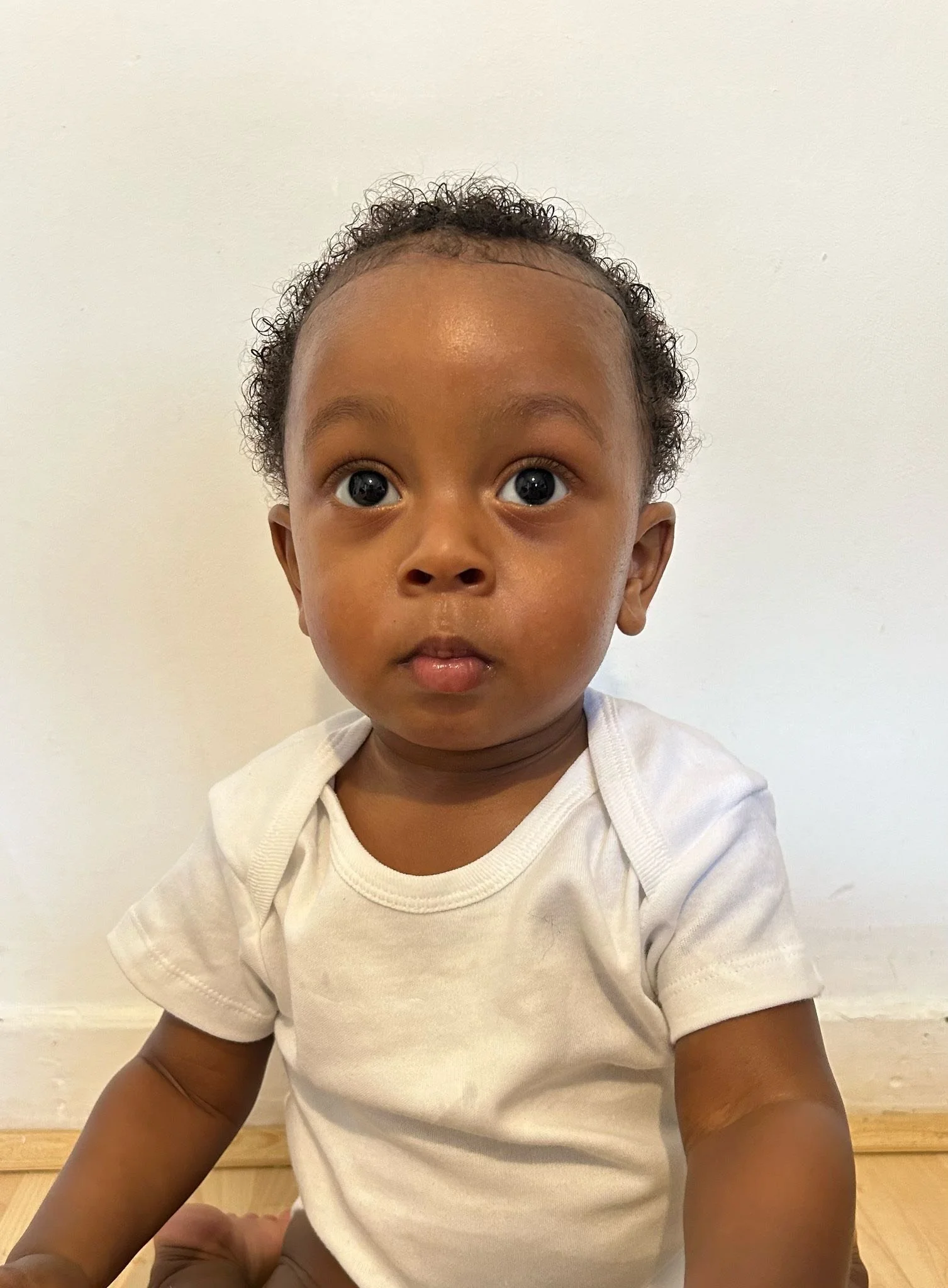 Close-up of a young child with big eyes and curly hair, sitting on a wooden floor against a white wall, wearing a white T-shirt. Modelling for leading child model and talent agency SocityUK based in London and Manchester.