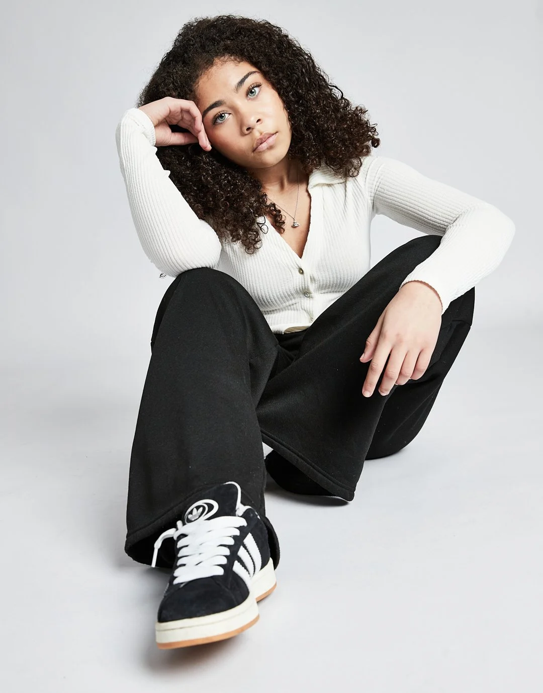 A young woman with curly hair sitting on the floor, wearing a white ribbed long-sleeve top, black wide-leg pants, and black sneakers with white stripes, resting her head on her hand.