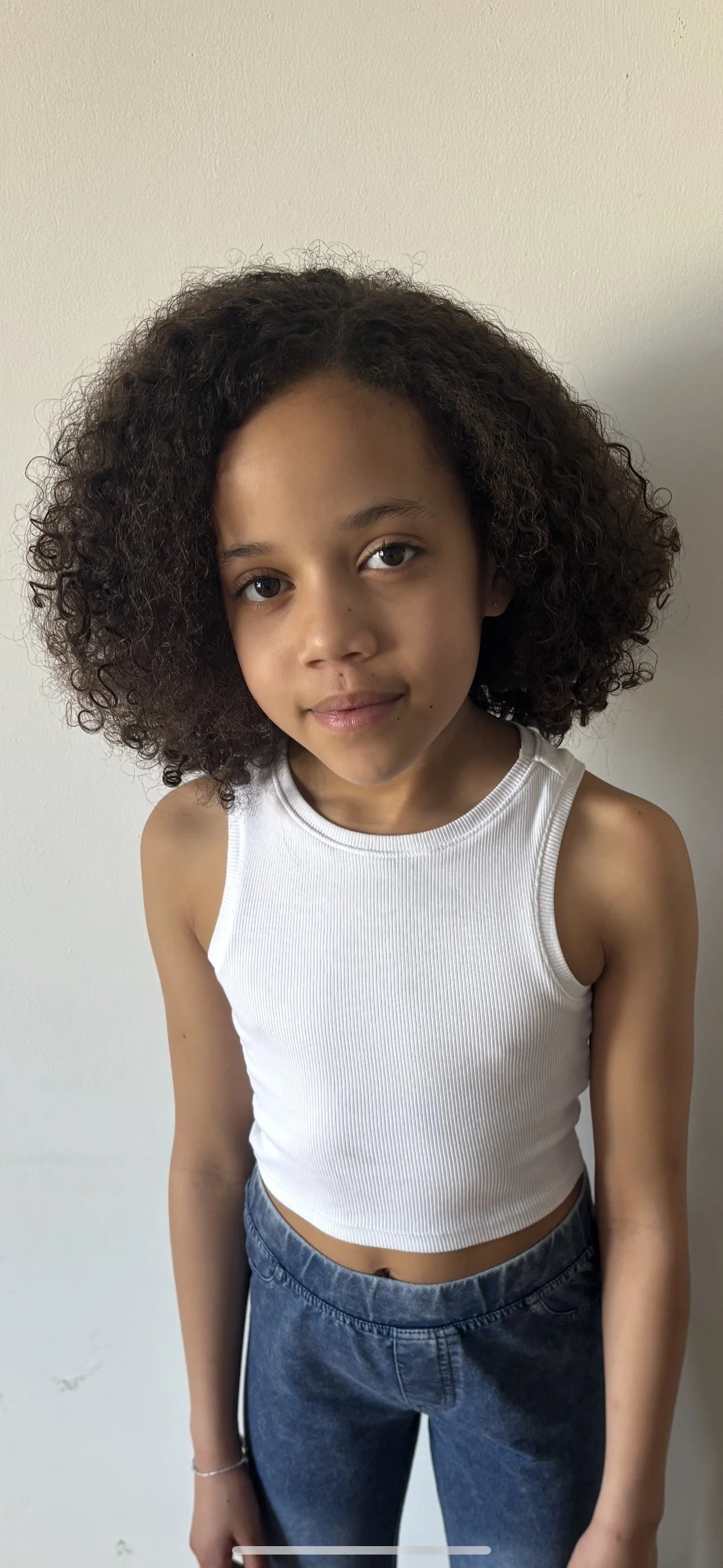 A young girl with curly dark hair and brown eyes, wearing a white sleeveless crop top and dark blue jeans, standing against a plain wall.