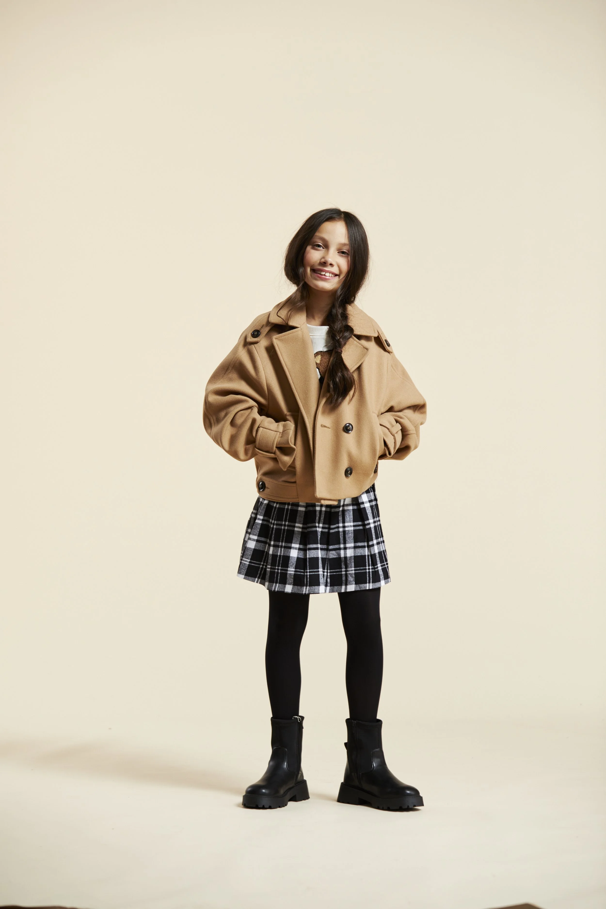A young girl smiling and standing with hands in pockets, wearing a tan jacket, plaid skirt, black tights, and black boots. Modelling for leading child model and talent agency SocityUK based in London and Manchester.