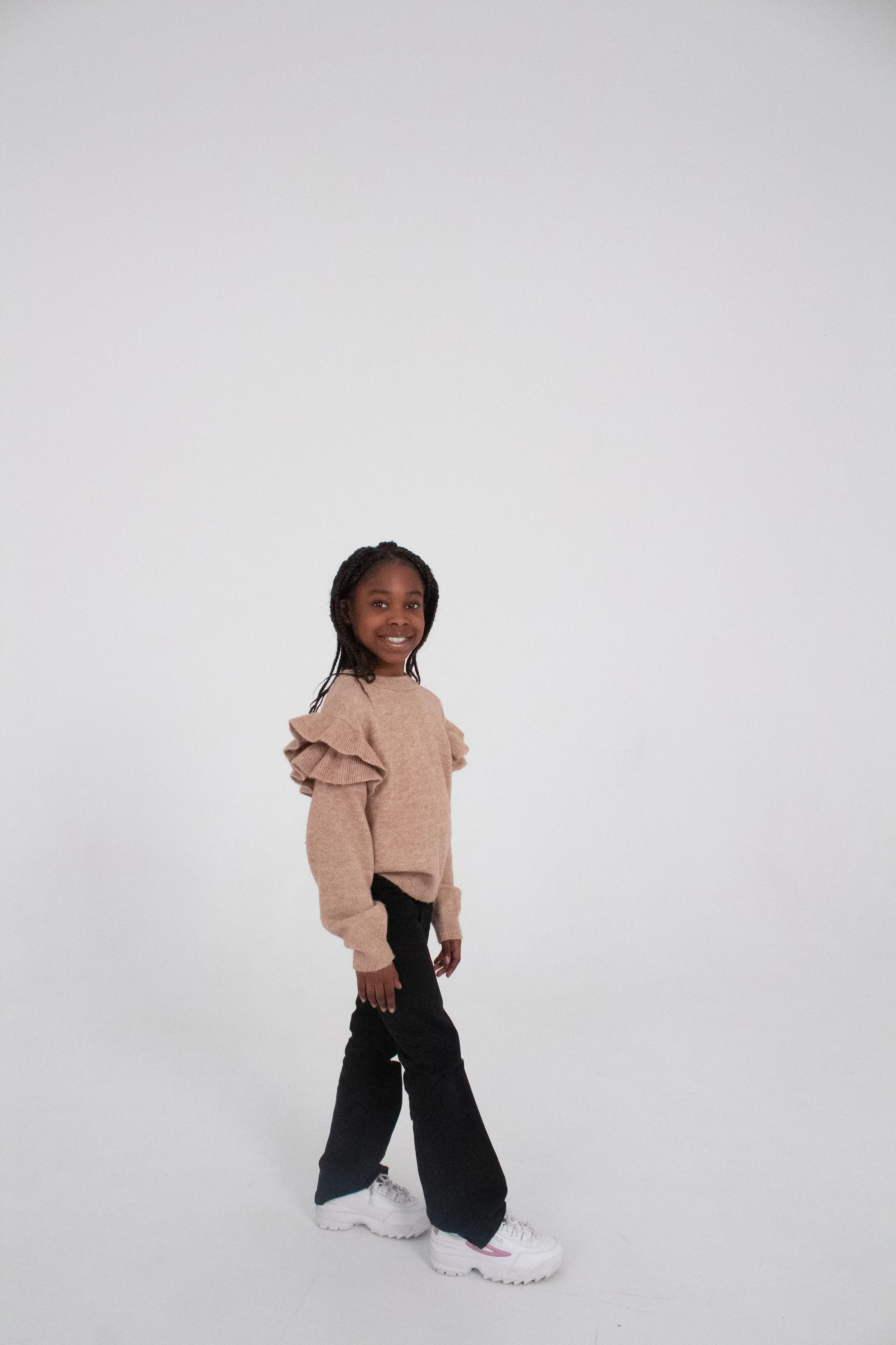A young girl smiling and standing in a plain, bright, white studio background. She is wearing a beige sweater with ruffled shoulders, black pants, and white chunky sneakers.