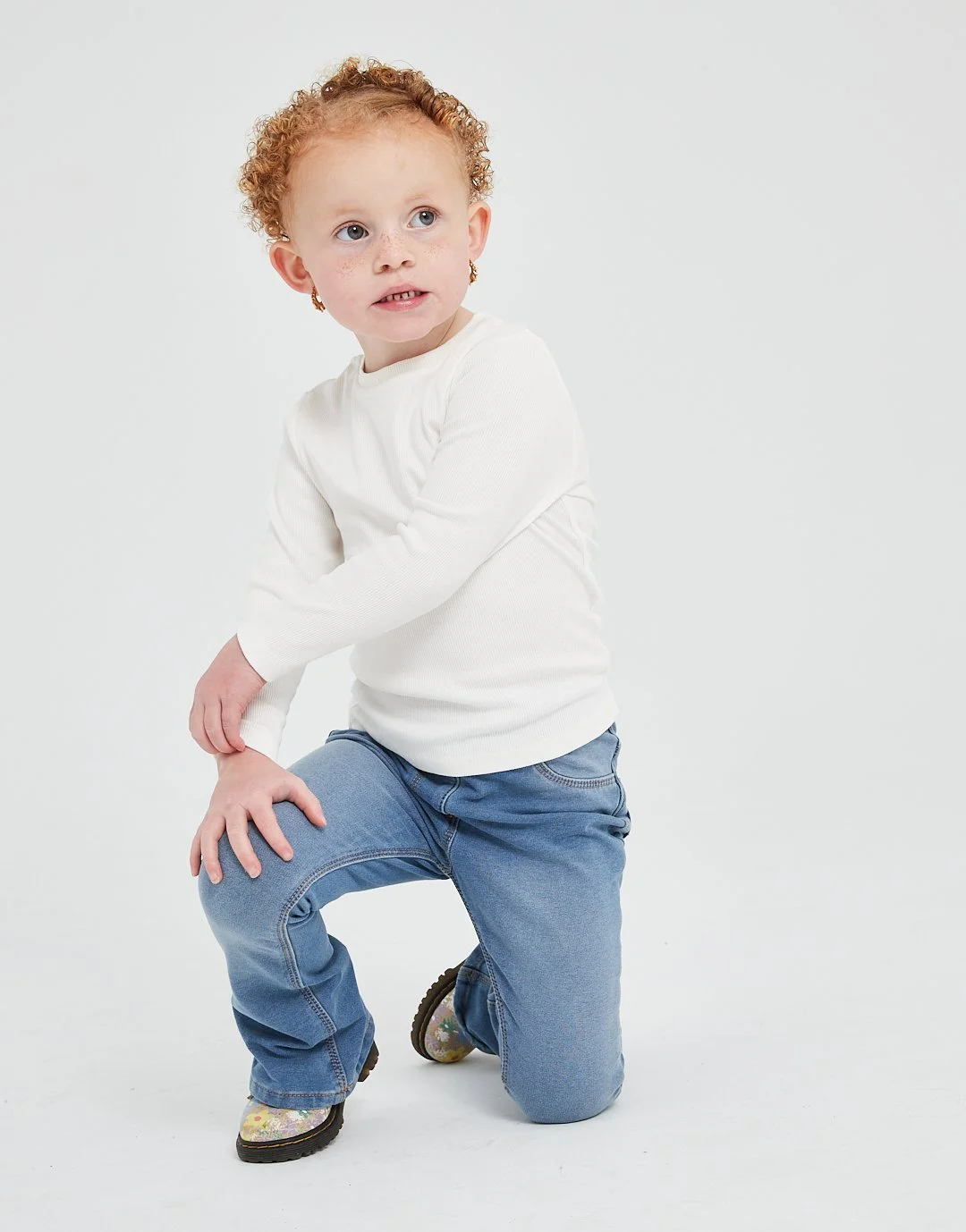 A young girl with curly red hair and freckles, wearing a white long-sleeve shirt, blue jeans, and yellow floral shoes, with one knee on the ground and her hand on her leg, looking slightly to the side against a plain white background.