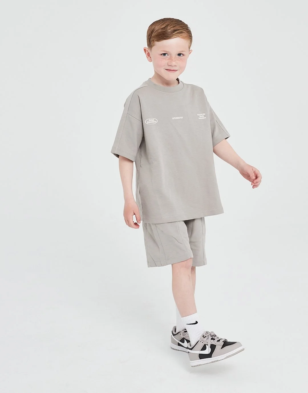 A young boy with red hair and freckles is walking against a plain white background, wearing a beige oversized T-shirt, beige shorts, white Nike socks, and beige and black sneakers. Modelling for leading child model and talent agency SocityUK based in