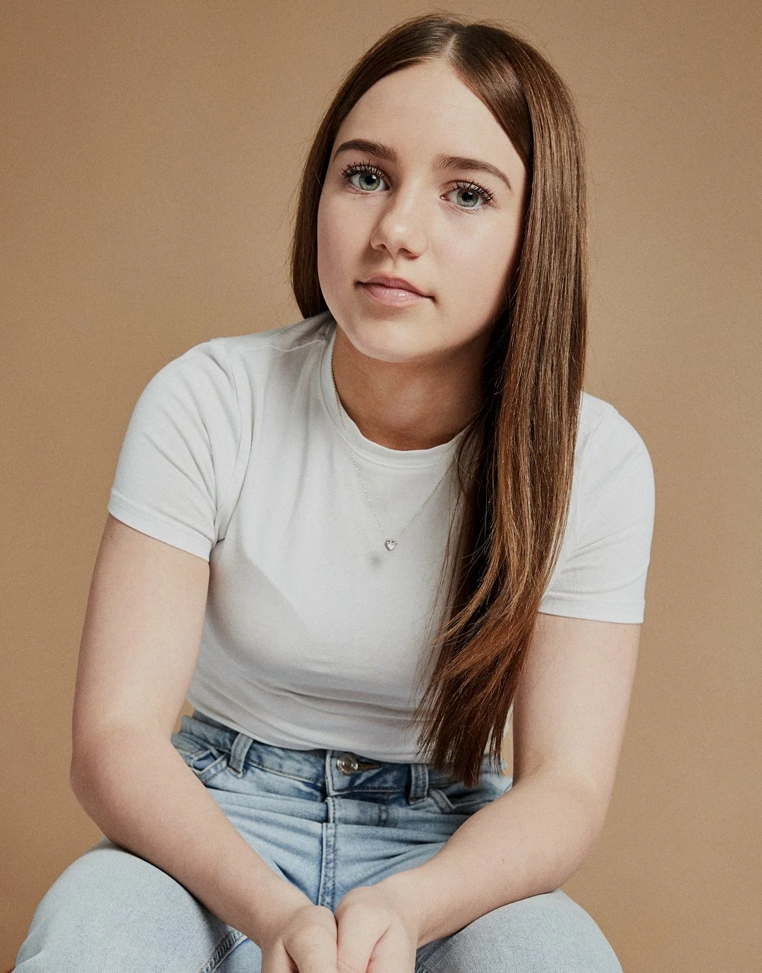 Young woman with long brown hair, wearing a white t-shirt and light blue jeans, sitting against a beige background.