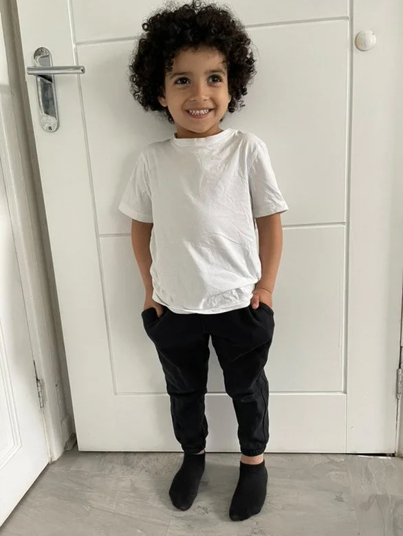 A young child with curly hair smiling and standing against a white door, wearing a white t-shirt, black pants, and black socks.