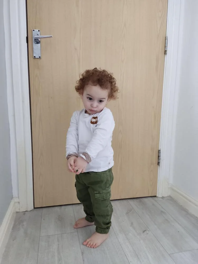 Young child with curly red hair standing barefoot in front of a wooden door indoors, wearing a white long-sleeve shirt and green pants.