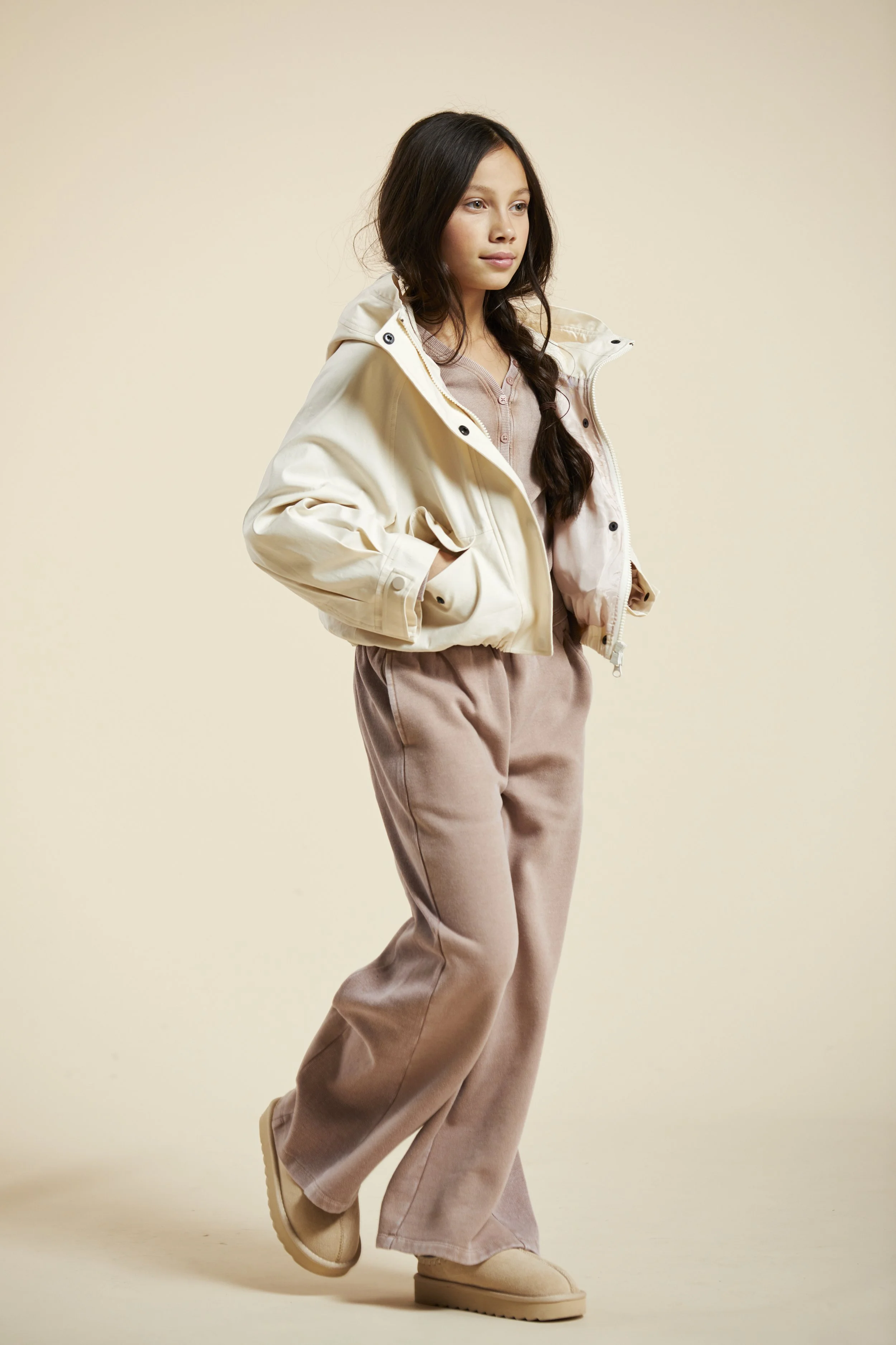 A young girl with long dark hair wearing a cream jacket, beige pants, and beige shoes, standing against a plain beige background. Modelling for leading child model and talent agency SocityUK based in London and Manchester.
