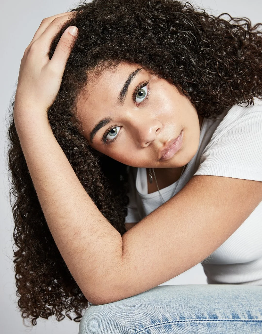 A young woman with curly hair and green eyes, wearing a white t-shirt, gazes directly at the camera with her head resting on one arm, posing against a neutral background.