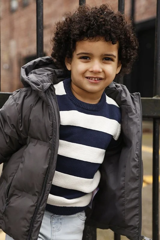 Young boy with curly hair smiling, wearing a black and white striped sweater, a dark gray puffy jacket, and light-colored pants, standing outdoors near a metal fence.