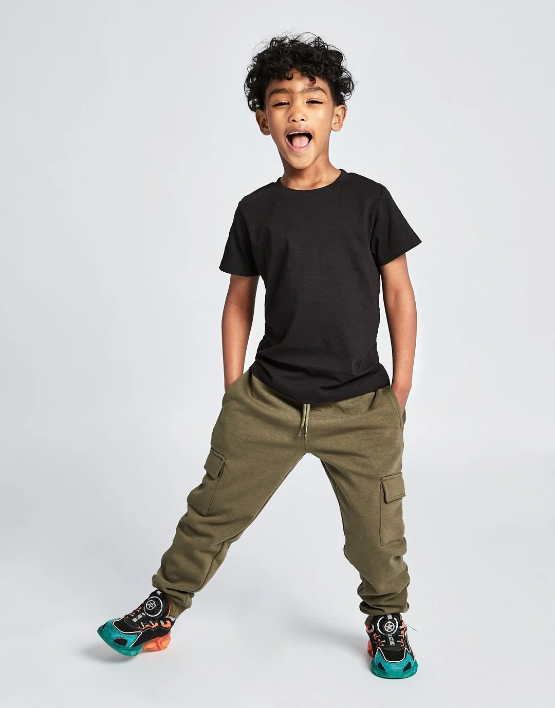 A young boy with curly black hair wearing a black t-shirt, green cargo pants, and colorful sneakers, standing with legs apart and hands in his pockets on a plain white background. Modelling for leading child model and talent agency SocityUK based in 