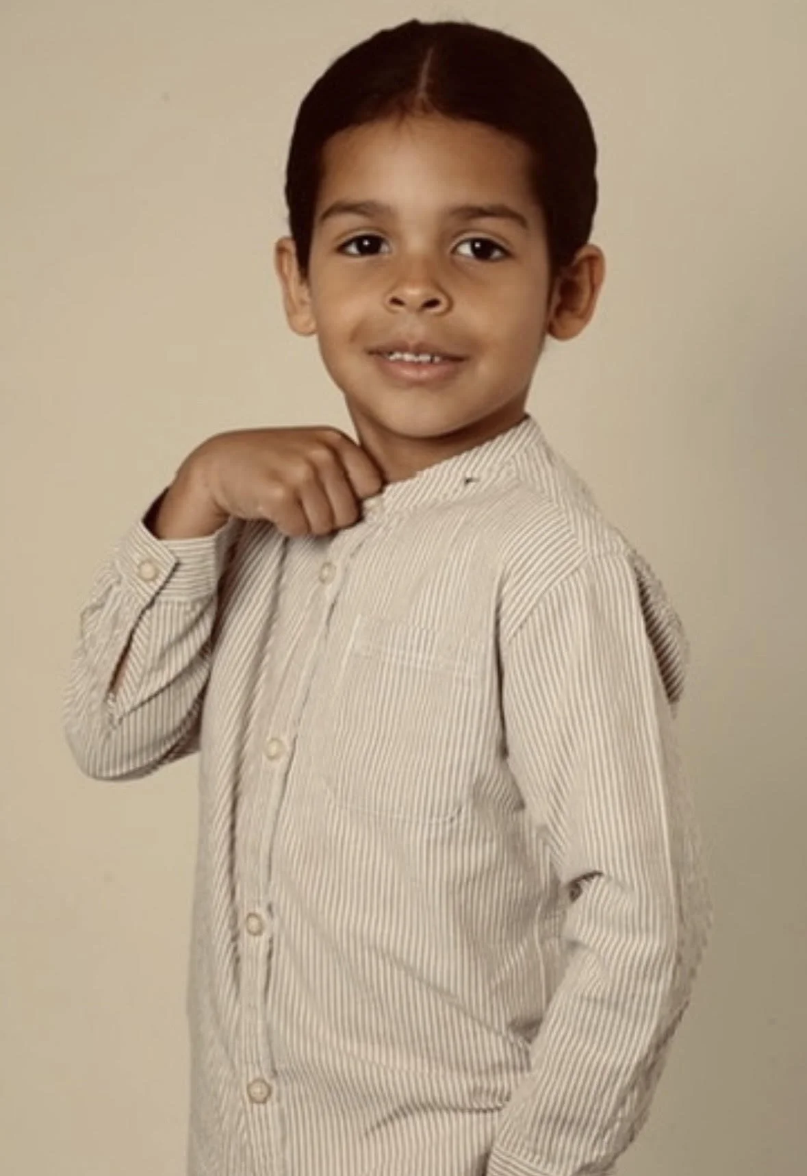 A young boy with dark hair, wearing a light-colored, striped button-up shirt, posing against a plain beige background.