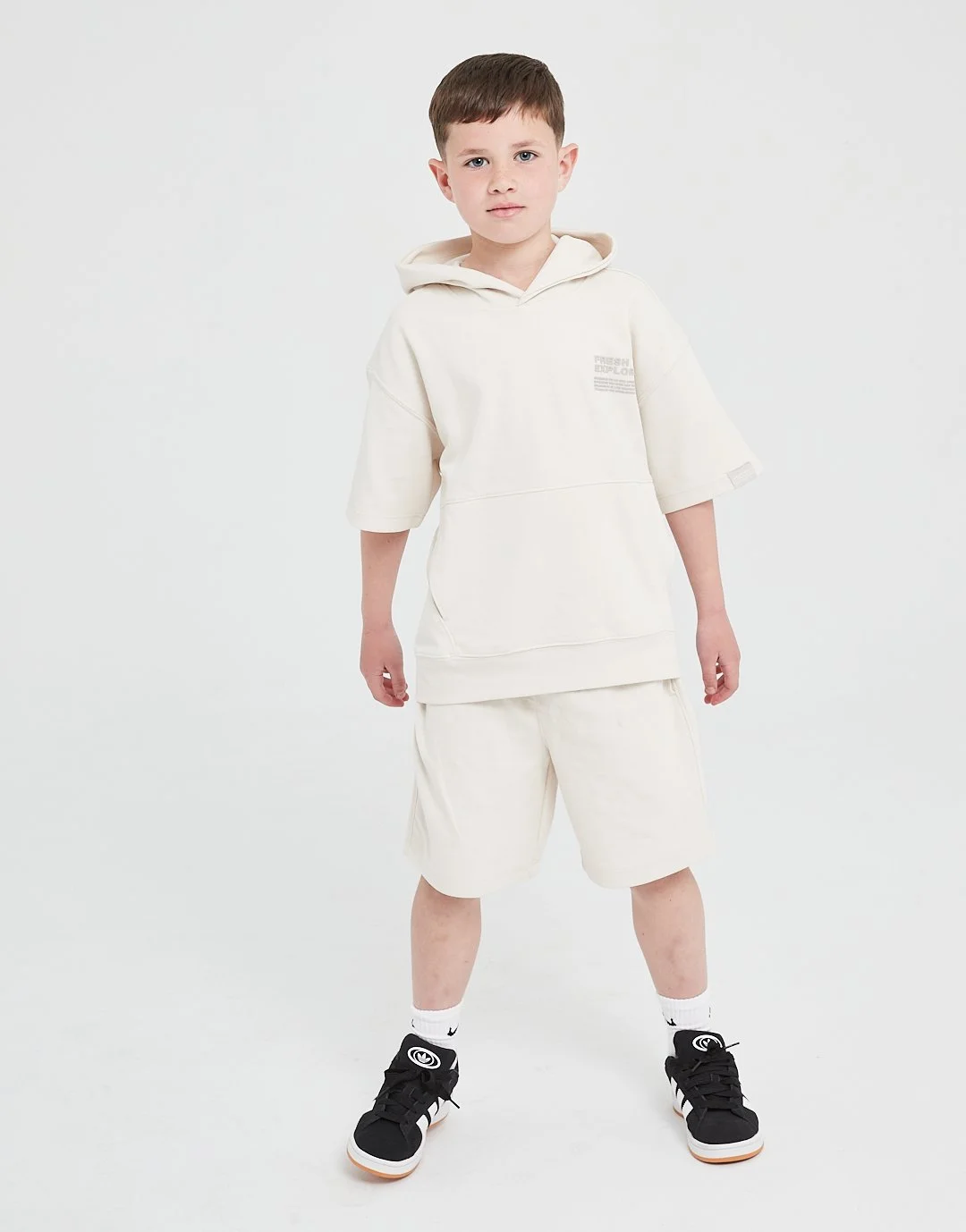 Young boy wearing an off-white hoodie, matching shorts, white socks, and black sneakers, standing against a plain white background.